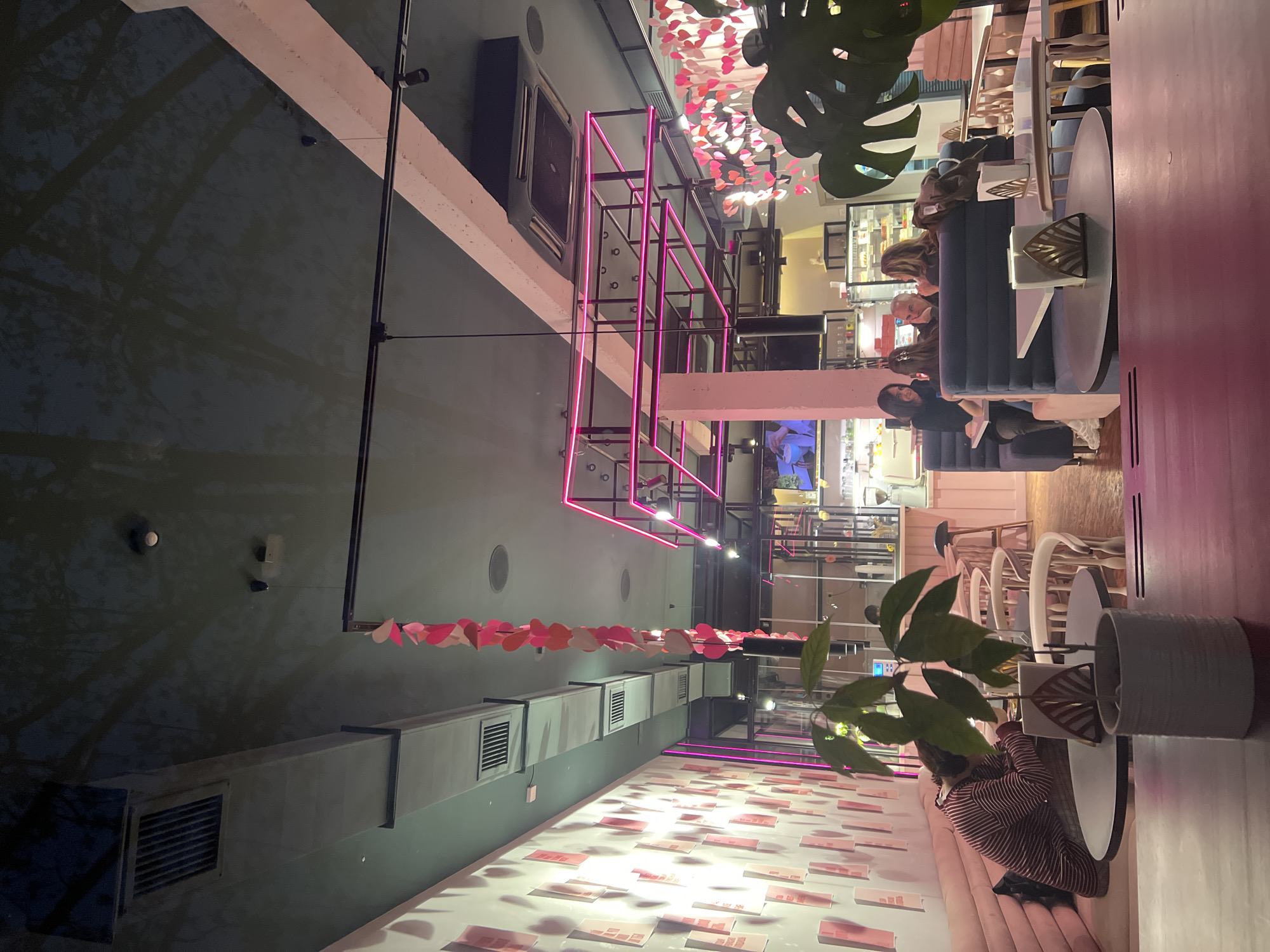 The image shows the interior of a restaurant in Krasnodar, Russia. The interior design features a pink and modern aesthetic with neon lighting and floral decorations. There are people seated at tables, some engaged in conversation or working, while others are waiting for their order. The restaurant has a variety of seating options, including booths and chairs. There are potted plants placed throughout the space. Overall, the atmosphere appears lively and stylish.