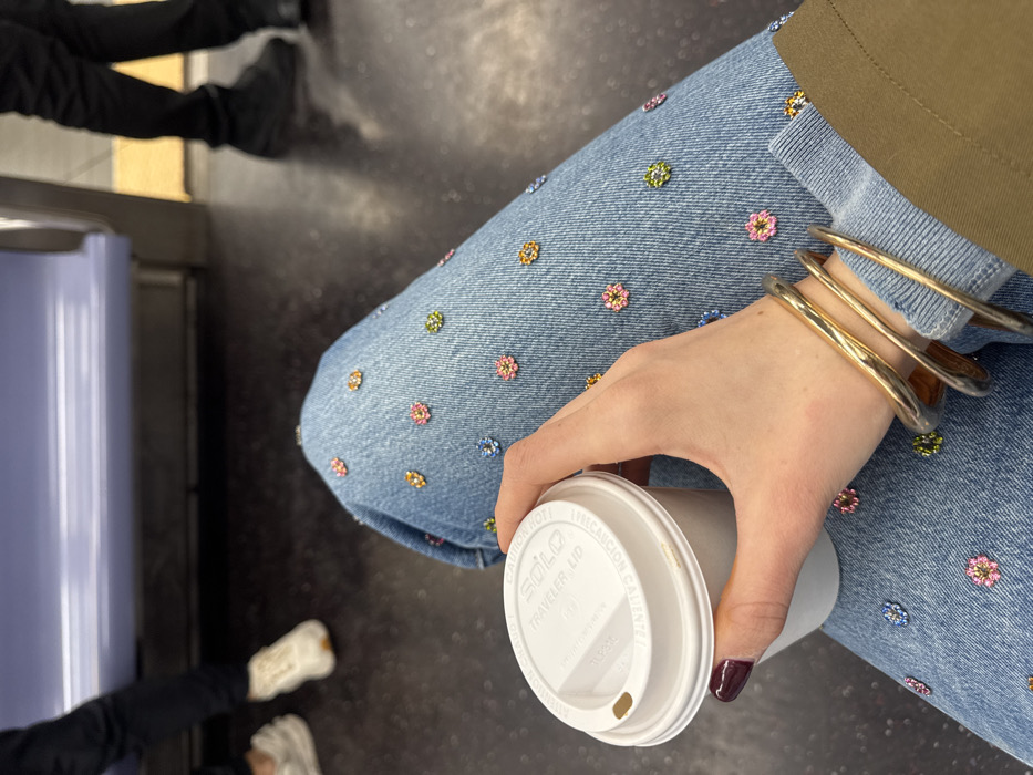 The image depicts an interior view of a public transit vehicle, identified as being in Hoboken, United States of America. The primary focus is the upper legs and right arm of a seated individual. This individual wears light blue denim jeans extensively adorned with small, multi-colored flower-shaped beads or appliqués. Their right hand, with dark red/burgundy fingernail polish, holds a white disposable coffee cup with a white "SOLO Traveler lid." Multiple gold or bronze bangles are visible on the wrist of the holding arm, and the cuff of a dark green or khaki sleeve is also present.
The floor of the vehicle is dark grey. In the background, to the left, the lower legs of another person wearing dark pants and white sneakers are partially visible. To the right, the lower legs and black shoes of a third individual, also in dark pants, are visible, standing near a yellow safety strip or line on the floor. A light purple/blue bench seat or railing is present in the mid-background.