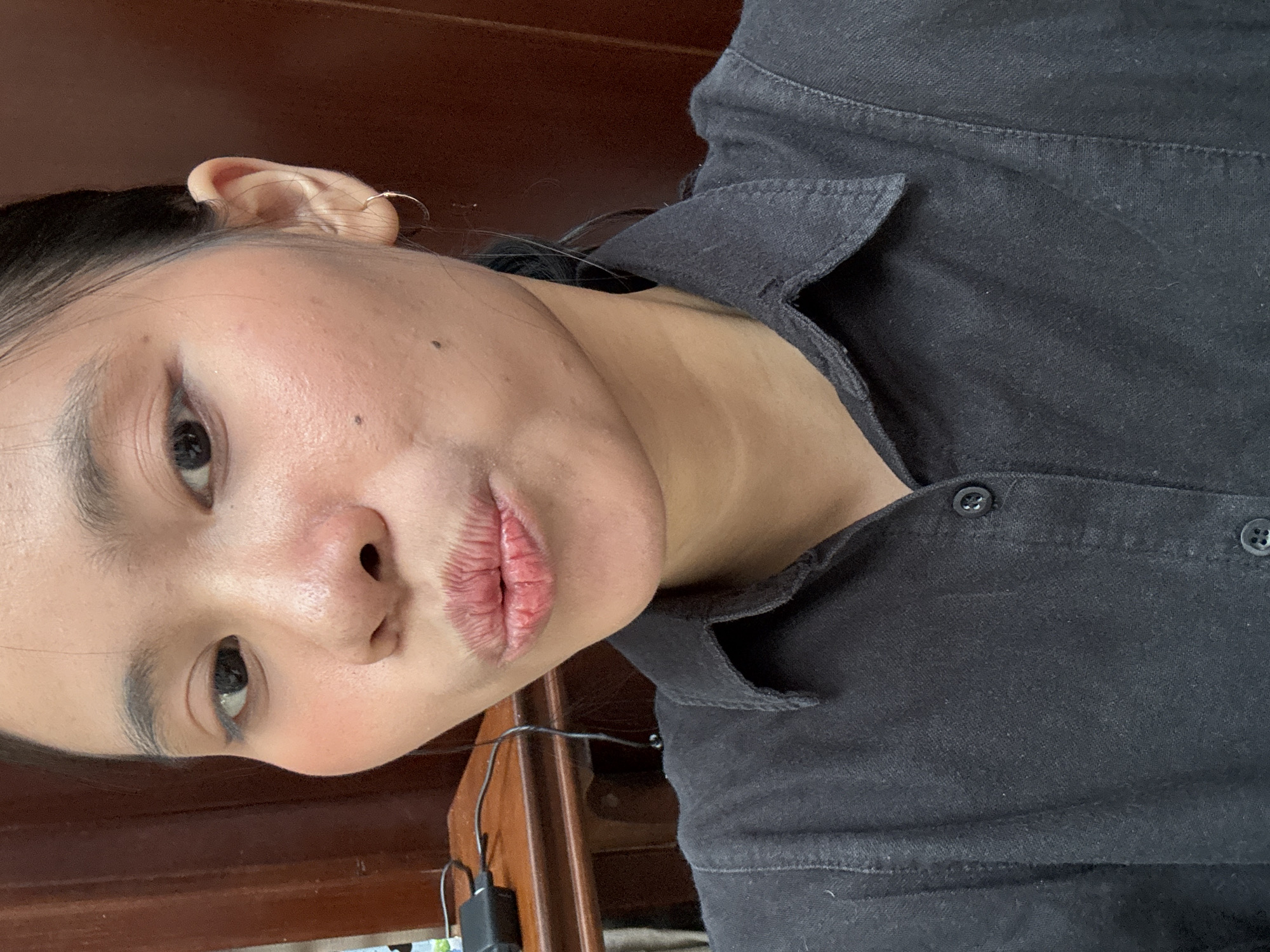 A medium shot shows a young woman in the foreground, her face tilted towards the camera with her lips pursed. She has dark hair pulled back, dark eyebrows, and wears a light application of makeup including blush and eyeliner. She has a small hoop earring in her visible right ear and two distinct moles on her right cheek. Her expression is playful. She is wearing a dark collared shirt, buttoned down the front. In the background, a wooden desk with a black electronic device and cables is visible to the left, and a dark brown wooden surface, possibly a door or wall, is to the right. The lighting suggests an indoor setting. The image was captured on August 21, 2022, at 10:37 AM WIB.