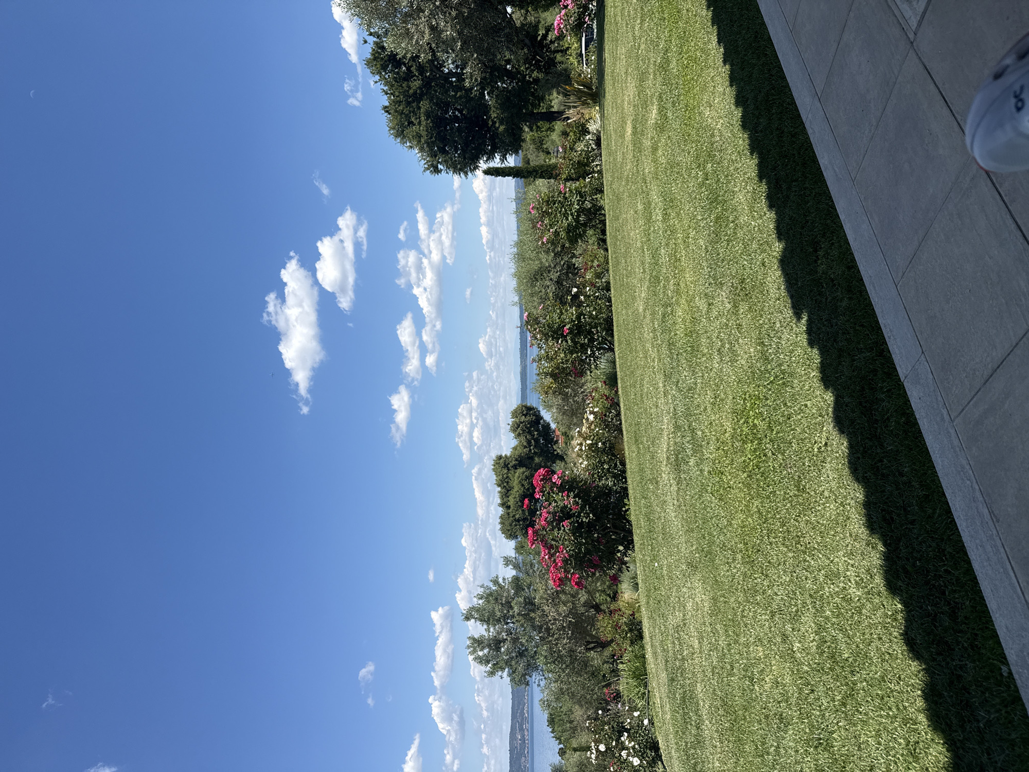 The scene depicts a view from a possible building or structure in Bracciano, Italy. A clear blue sky with scattered cumulus clouds dominates the left portion of the image, with a possible faint moon in the upper left corner. A body of water, likely Lake Bracciano, is visible in the middle distance. The right portion shows a well-maintained green lawn. Flower beds and trees separate the lawn from the distant landscape. A grey tiled area suggests a patio or walkway adjoining the lawn. A partially visible white shoe is present on the rightmost edge of the image.