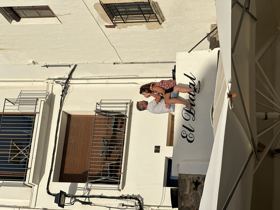 The image depicts a sunlit outdoor scene in Calella De Palafrugell, Spain, featuring two individuals engaged in a close interaction.
**Location and Environment:** The scene is set on a narrow thoroughfare between two multi-story, white-plastered buildings. Strong sunlight from the right casts distinct shadows. In the immediate foreground, the top portions of two large, white outdoor umbrellas are visible, indicating a café or restaurant setting below.
**Midground Subjects and Objects:** A Caucasian male and female, appearing to be middle-aged, are seated side-by-side on a low, white plastered wall. The wall bears the black script lettering "El Didal." The man is on the left, wearing a light-colored, possibly white or light grey, short-sleeved patterned shirt, blue shorts, and light brown loafers. He has short, greying hair and a short grey beard. His right arm is around the woman's lower back, and his left hand is cupping her cheek/jaw. The woman, on the right, wears a red, white, and black patterned short-sleeved top or dress, dark shorts, and dark patterned flat shoes. She has curly brown hair. Her right hand is raised, covering her mouth and chin, while her left arm is around the man's waist. They are positioned facing each other, engaged in what appears to be an intimate or affectionate moment, possibly laughing or talking closely. Behind them, on the building wall, a dark number "7" is affixed.
**Background Architecture:**
* **Left Building:** Features multiple balconies with metal railings. The lowest visible balcony has dark brown louvered shutters that are closed, behind which two dark chairs and a small table are arranged. Above this, another balcony with a similar railing shows one chair and a table. A black streetlamp fixture with a curved arm is mounted on the wall, accompanied by numerous black electrical conduits and wires running vertically and horizontally. Below the wall where the couple sits, a stone base and a dark blue container are visible.
* **Right Building:** Features a window with closed dark brown louvered shutters in the upper section and a grated window below it. A small, red security alarm box is visible on the wall near the top window. Electrical conduits and pipes are also visible along the white facade.