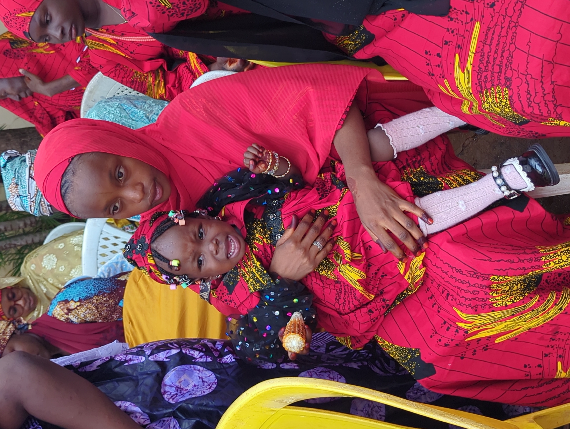 The image captures a lively social gathering, likely taking place in Unguwar Rimi, Nigeria. The central focus is a young woman holding a small child on her lap. The young woman wears a red headscarf and a vibrant red dress patterned with black and yellow designs. She looks subtly upwards with a calm expression, her arm gently around the child.
The small child, likely a girl, mirrors the woman's attire with a matching red patterned dress featuring black ruffled sleeves. Her braided hair is adorned with colorful beads. She has a wide, joyful smile, showing her teeth, as she looks directly at the camera. In her right hand, she holds a partially consumed piece of corn on the cob. Her feet are clad in white ruffled socks and black Mary Jane-style shoes embellished with pearls. The woman's hand displays a ring and bangles.
Surrounding these two central figures are several other individuals, mostly women, also dressed in an array of colorful traditional fabrics, including shades of red, gold, blue, and purple. A white plastic chair is partially visible in the background, suggesting an informal indoor or shaded outdoor setting. The scene conveys a sense of warmth and communal happiness, underscored by the child's cheerful demeanor. The image appears to be taken during the daytime. No text is visible.