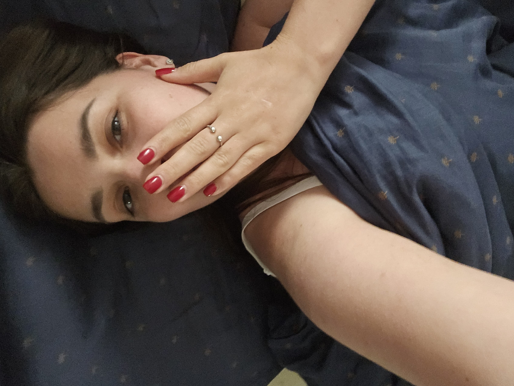 The image shows a close-up of a woman lying in bed, with her right arm and hand covering her mouth. She has bright red nail polish on her fingernails and is wearing a ring on her left hand. She is wearing a white bra strap visible. The bedding is a dark blue fabric with a subtle pattern of small, gold-colored trees. The background is dark and out of focus, suggesting an indoor setting. The image does not contain any information or context related to the city of Petropavlivska Borshchahivka, Ukraine.