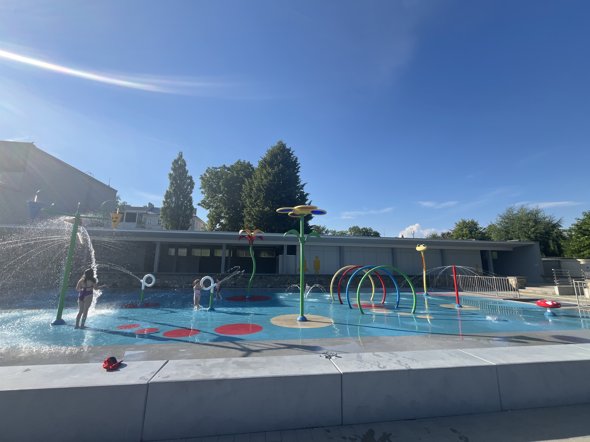 This is a wide shot of an empty splash pad in a park on a sunny day. In the foreground, there is a gray concrete ledge. The splash pad is bright blue with red and yellow circular designs on the surface. There are several water features, including a tall green pole with a flower-like top, a yellow frog-shaped spout, and rainbow-colored arches that spray water. In the background, there is a long, low building with a gray roof and windows. There are trees behind the building, and a clear blue sky with some wispy clouds. There are no people in the image, but there is a red hat on the concrete ledge in the foreground. The scene is well-lit and appears to be clean and well-maintained.