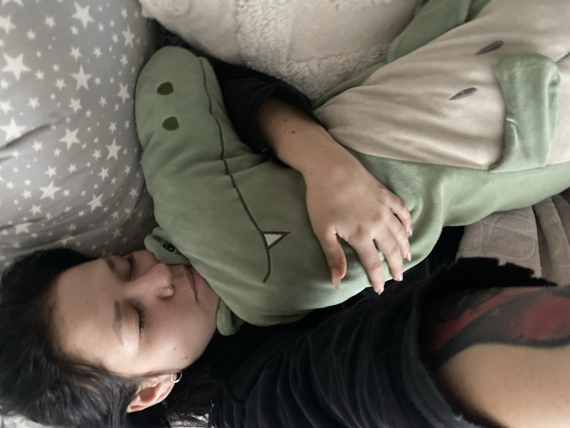A person is lying in bed with a pillow featuring a star pattern to their left. They are wearing a black shirt and a dinosaur onesie. Part of a tattoo is visible on their right arm. The location is unknown and cannot be determined from the image.