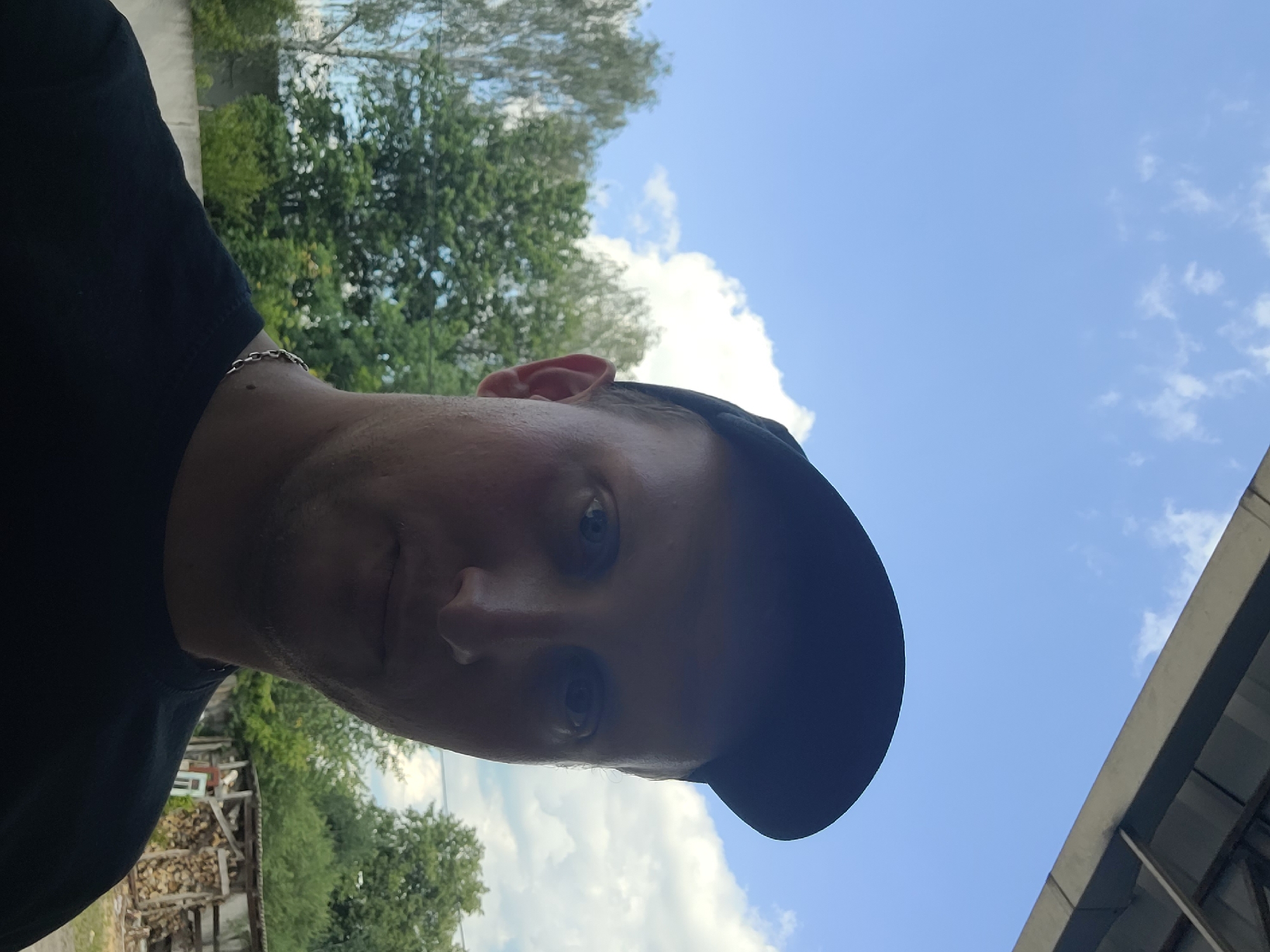 A man is taking a selfie outdoors. He is wearing a black t-shirt and a black baseball cap. His head is tilted to the side. The background shows a blue sky with white clouds, green trees, and a structure with stacked wood. The lighting suggests it is daytime.