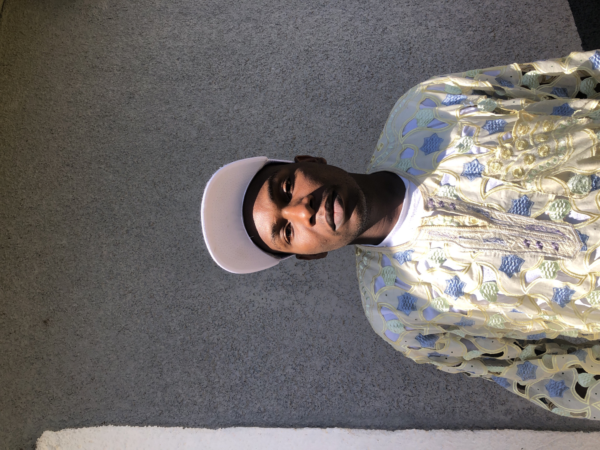 A medium shot captures a man standing in front of a textured grey wall. He is wearing a white baseball cap and a light-colored, intricate garment with cutouts and embroidered patterns in shades of blue, green, and yellow. The man is looking directly at the camera, with sunlight casting shadows on his face, highlighting his features. To the left of the man, a portion of a white, textured pillar is visible. The overall setting suggests an outdoor location, possibly in the city of Jos, Nigeria, given the architectural style of the wall and pillar.