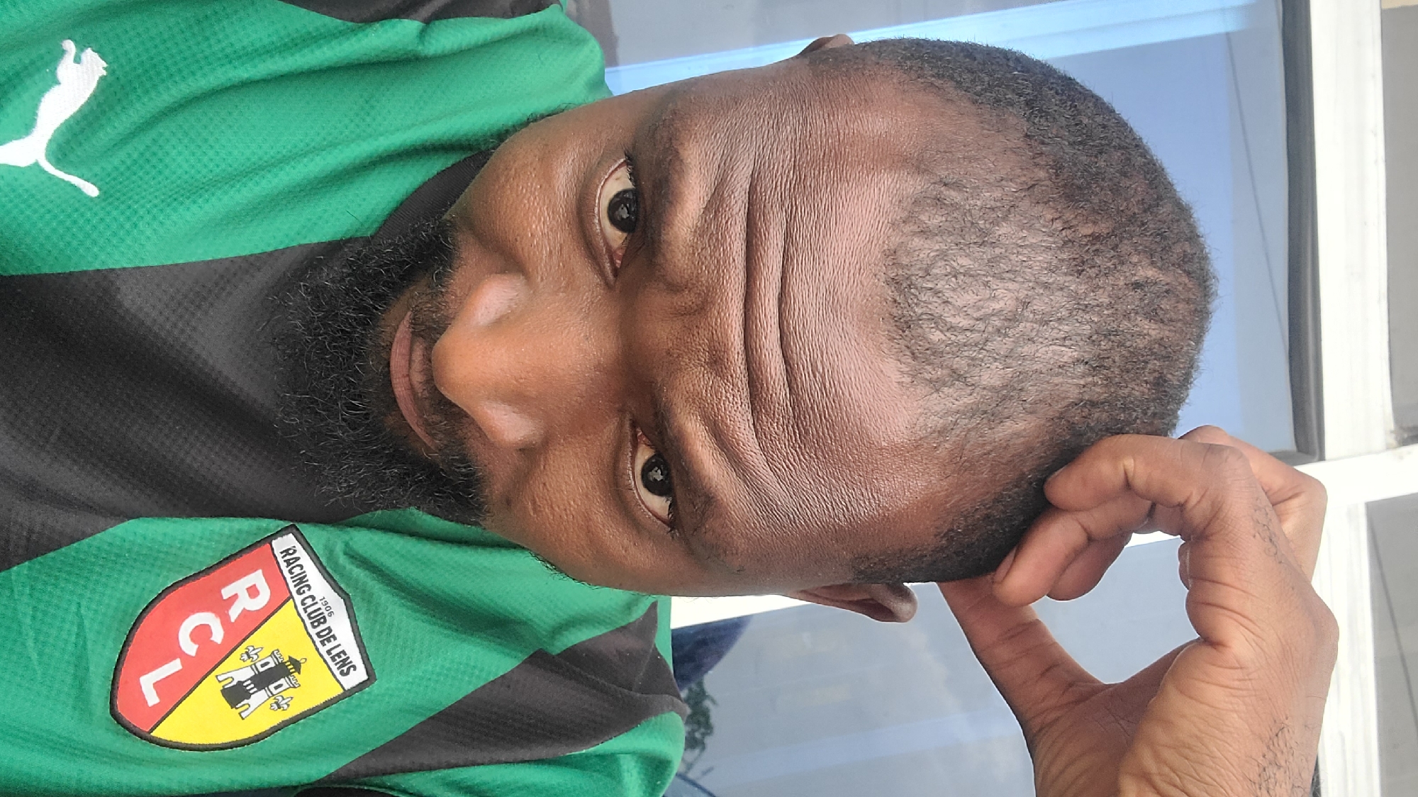 A close-up, Dutch-angle portrait captures a dark-skinned man with a full, dark beard and short, curly hair. He is wearing a green and black striped or paneled sports jersey. On the upper green portion of the jersey, a white Puma logo is visible. Below, on the left chest, is the emblem of Racing Club de Lens, a French football club. The badge is red, yellow, black, and white, and clearly displays the text "RACING CLUB DE LENS", "RCL", and the year "1906".
The man looks directly at the camera with a direct gaze. His forehead shows prominent horizontal wrinkles, suggesting an expression of concentration, contemplation, or possibly fatigue. His right hand is raised, with fingers gently pressing against the side of his head near his temple or ear. The background is indistinct, appearing as a soft blend of light blue and white, potentially from a window or light-colored wall. The image is well-lit, likely by natural daylight. This scene is set in Jos, Nigeria, showing the man in a reflective or focused moment while displaying his affiliation with the Racing Club de Lens.