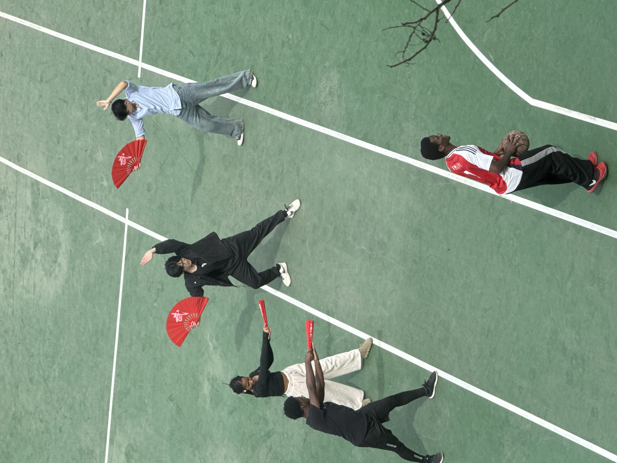 The image depicts five individuals on an outdoor, light-green sports court marked with white lines, located in Daxing District, China.
In the upper-right section, two light-skinned males are engaged in a performance or practice. One male wears a light blue button-down shirt, blue jeans, and white sneakers; he holds an open red fan emblazoned with white Chinese characters (likely '武') in his left hand, his right hand raised. The second male, dressed in a black jacket, black pants, and white sneakers, similarly holds a red fan with white Chinese characters in his left hand, striking a dynamic pose.
Positioned centrally on the left, a dark-skinned female, wearing a black long-sleeved crop top, light-colored loose pants, and light-colored shoes, holds two red cylindrical batons extended forward with both hands. Adjacent to her, a dark-skinned male, wearing a black t-shirt, black shorts over leggings, and black sneakers, mirrors her stance, also holding two red cylindrical batons extended forward.
Separated from the performance group and positioned in the lower-right section, a dark-skinned male, wearing a red and white short-sleeved jersey with the number "1" partially visible, black track pants with white stripes, and red sneakers, holds a brown basketball with both hands. He is looking towards his left.
A bare tree branch extends into the frame from the far right side, near the basketball player. The court surface appears slightly worn in areas.