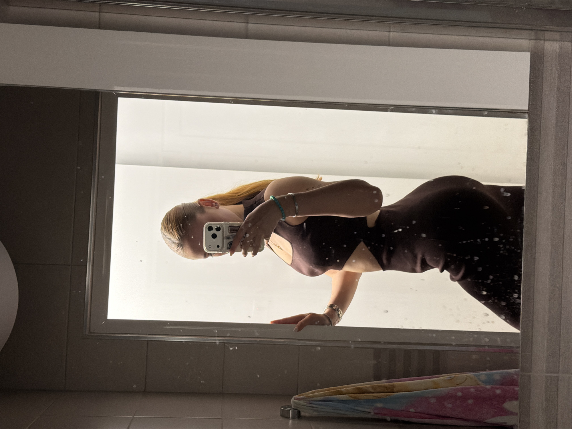 A woman is reflected in a bathroom mirror. She is wearing a dark brown dress with a cut-out design. She is holding a mobile phone, presumably taking the picture. The background is blurred and brightly lit, likely a shower or bathroom area. An umbrella is visible in the bottom corner of the mirror's reflection. The location is undetermined and not possible to establish from the image. The image doesn't feature any information indicating it was taken in Plovdiv, Bulgaria.