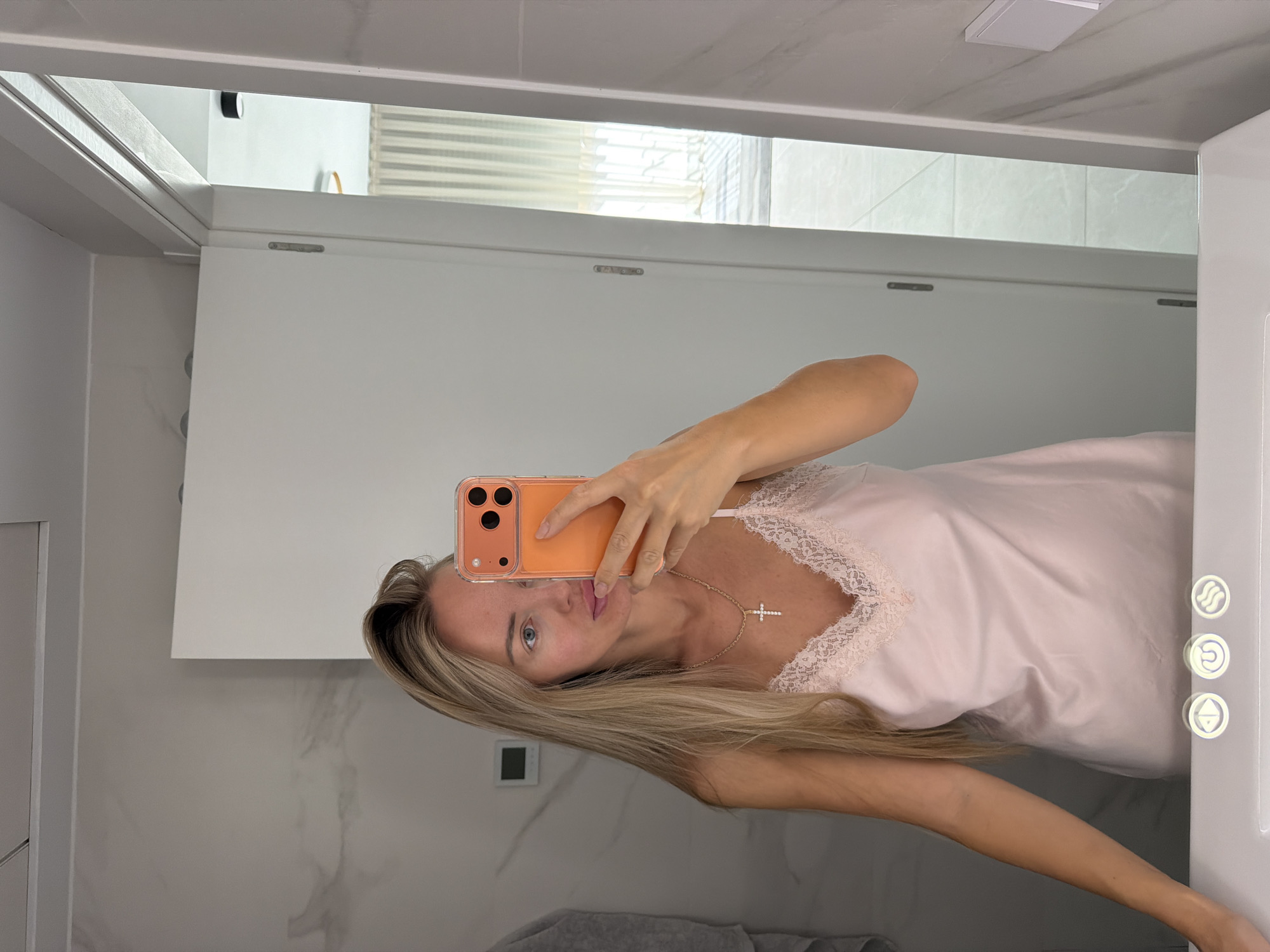 A woman is taking a mirror selfie in a modern bathroom. She is wearing a pale pink slip dress with lace trim and a gold cross necklace. Her blonde hair is long and straight. She is holding an orange smartphone in her right hand, which is angled to capture her reflection. The bathroom features marble-patterned walls, a white door, and a white vanity with a sink. In the background, through an open doorway, a sunlit room with sheer curtains is visible. The location is identified as Finestrat, Spain.
