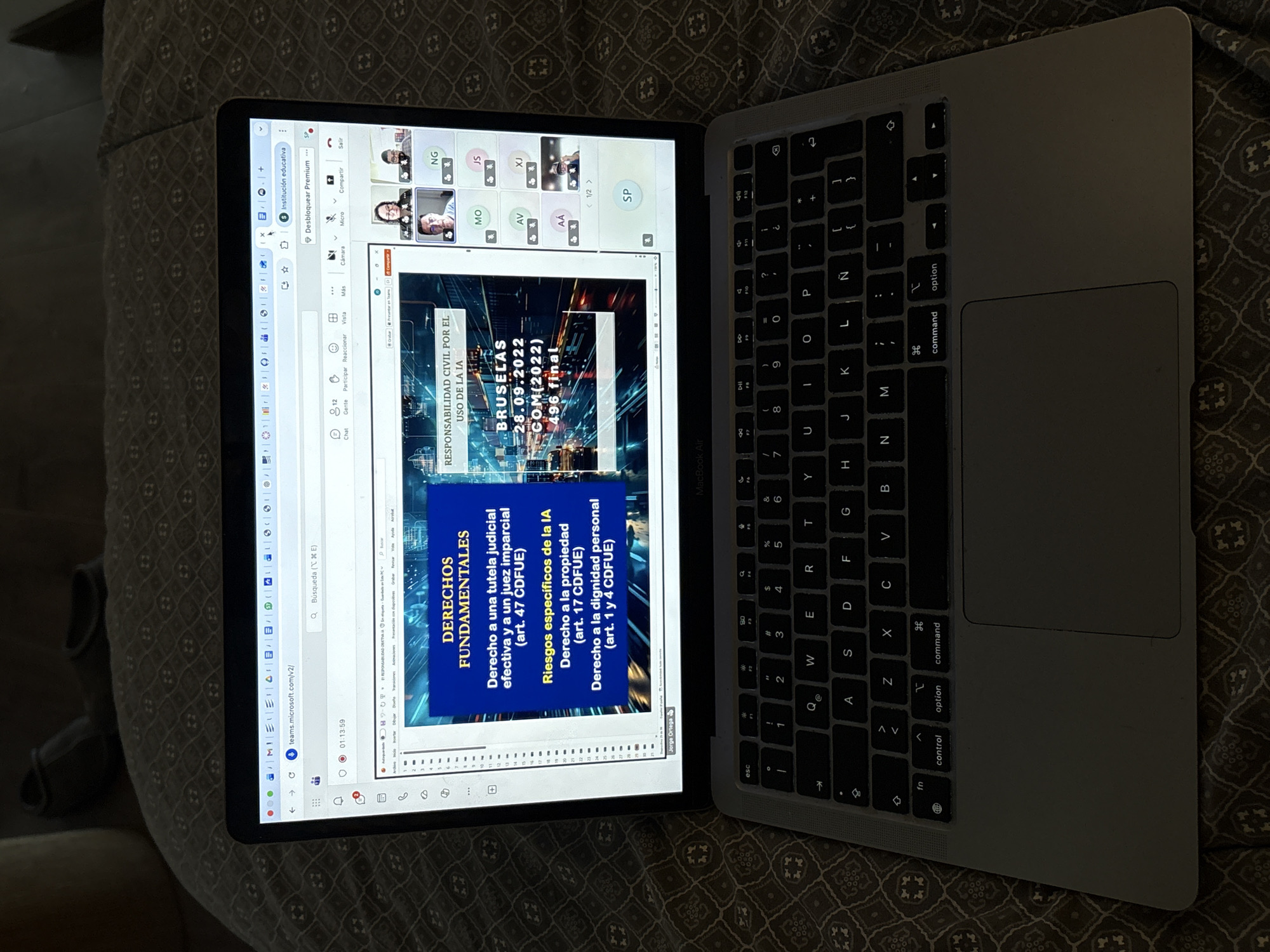 An overhead shot captures a MacBook Air laptop resting on a dark patterned blanket or duvet in San Bernardino Tlaxcalancingo, Mexico. The laptop screen displays an active Microsoft Teams meeting. The main content on the screen is a presentation slide titled "RESPONSABILIDAD CIVIL POR EL USO DE LA IA" (Civil Liability for the Use of AI). The slide further details "DERECHOS FUNDAMENTALES" (Fundamental Rights), listing points such as "Derecho a una tutela judicial efectiva y a un juez imparcial (art. 47 CDFUE)," "Riesgos específicos de la IA," "Derecho a la propiedad (art. 17 CDFUE)," and "Derecho a la dignidad personal (art. 1 y 4 CDFUE)." Event details "BRUSELAS," "28.09.2022," and "COM(2022) 496 final" are also visible on the slide.
At the top right of the screen, several participants are seen in small video feeds, identified by initials like NG, JS, MO, AV, XJ, AA, and SP. The Teams interface shows various controls, including "Chat," "Gente" (People), "Participar" (Participate), "Reaccionar" (React), "Cámara" (Camera), "Micro" (Mic), "Compartir" (Share), and "Salir" (Exit). A timestamp "01:13:59" is visible on the browser tab, which also shows "teams.microsoft.com/v2/". The name "Jorge Ortega" is faintly visible at the bottom left, likely indicating the presenter. A person's leg, covered by a light-colored fabric, is partially visible in the background on the left. The overall lighting is dim, suggesting an indoor setting during the evening or night.