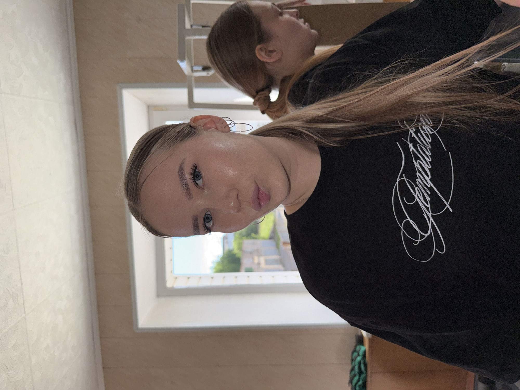 A young woman with blonde hair pulled into a ponytail, wearing a black t-shirt with white lettering, poses for a selfie in a room. She has on hoop earrings and is wearing makeup. Her face is turned slightly to her left, and her lips are pursed. In the background, a window shows an outdoor scene with buildings and trees. Another young woman with blonde hair in a ponytail is visible in a mirror behind her, looking down. The room appears to be domestic, with pale walls and a textured ceiling. The overall lighting suggests it is daytime.