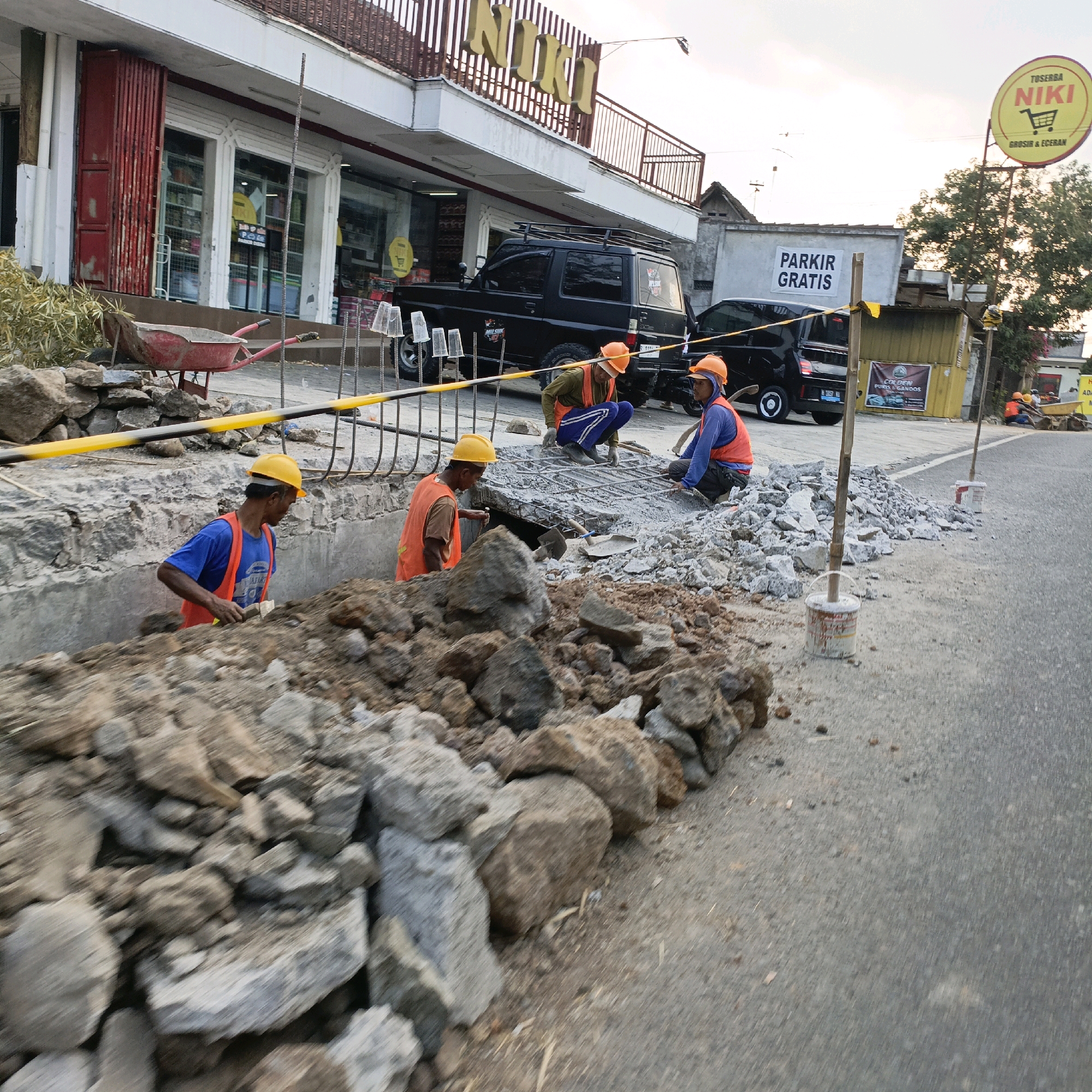 The image depicts a street scene in Salatiga, Indonesia, where construction work is underway. Several construction workers, wearing orange safety vests and hard hats, are engaged in activities related to road or building repair. One worker in the foreground, wearing a blue shirt and orange vest, is holding a shovel and appears to be working in a dug-out area. In the middle ground, two other workers are crouched over a section of rebar and concrete, possibly working on reinforcing a structure.
Behind the construction site, a black SUV is parked, and further back, a black van is visible. A building with the prominent sign "NIKI" is situated to the left, appearing to be a store or commercial establishment. A yellow sign advertising "PARKIR GRATIS" (Free Parking) is also visible on a building to the right. The environment suggests an urban or semi-urban setting, with a road surface, piles of construction debris including rocks and concrete, and vegetation in the background. The sky is overcast, indicating potential for changing weather conditions. The overall scene captures a moment of active construction and daily life in Salatiga.