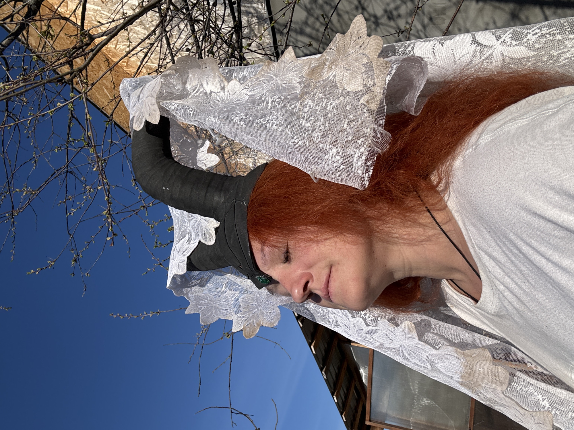 The image shows a person with red hair wearing a white dress, a lace veil and a black horned helmet. The person is outside. Behind the person are branches of a tree, a blue sky, a piece of plywood, and a building. It is daytime. The photo was taken in Chaikovsky, Russia.