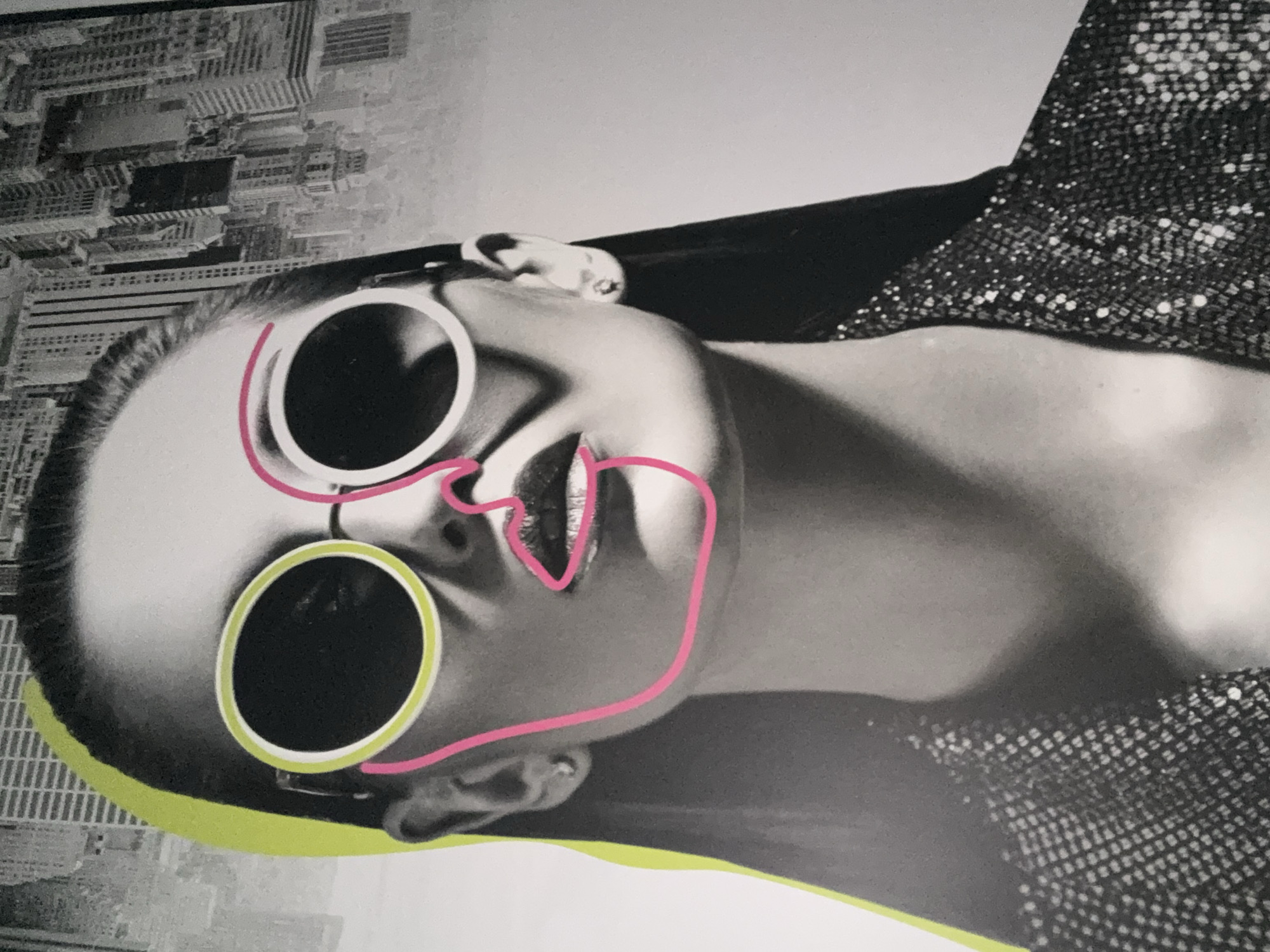 The image is a black and white photograph of a woman's face, partially overlaid with bright pink and lime green lines. The woman is wearing large, round sunglasses with white and lime green rims. Her face is angled to the side, and the pink lines accentuate her lips and jawline. In the background, there is a blurred image of a cityscape, with tall buildings visible. The woman is wearing a dark, sequined jacket.