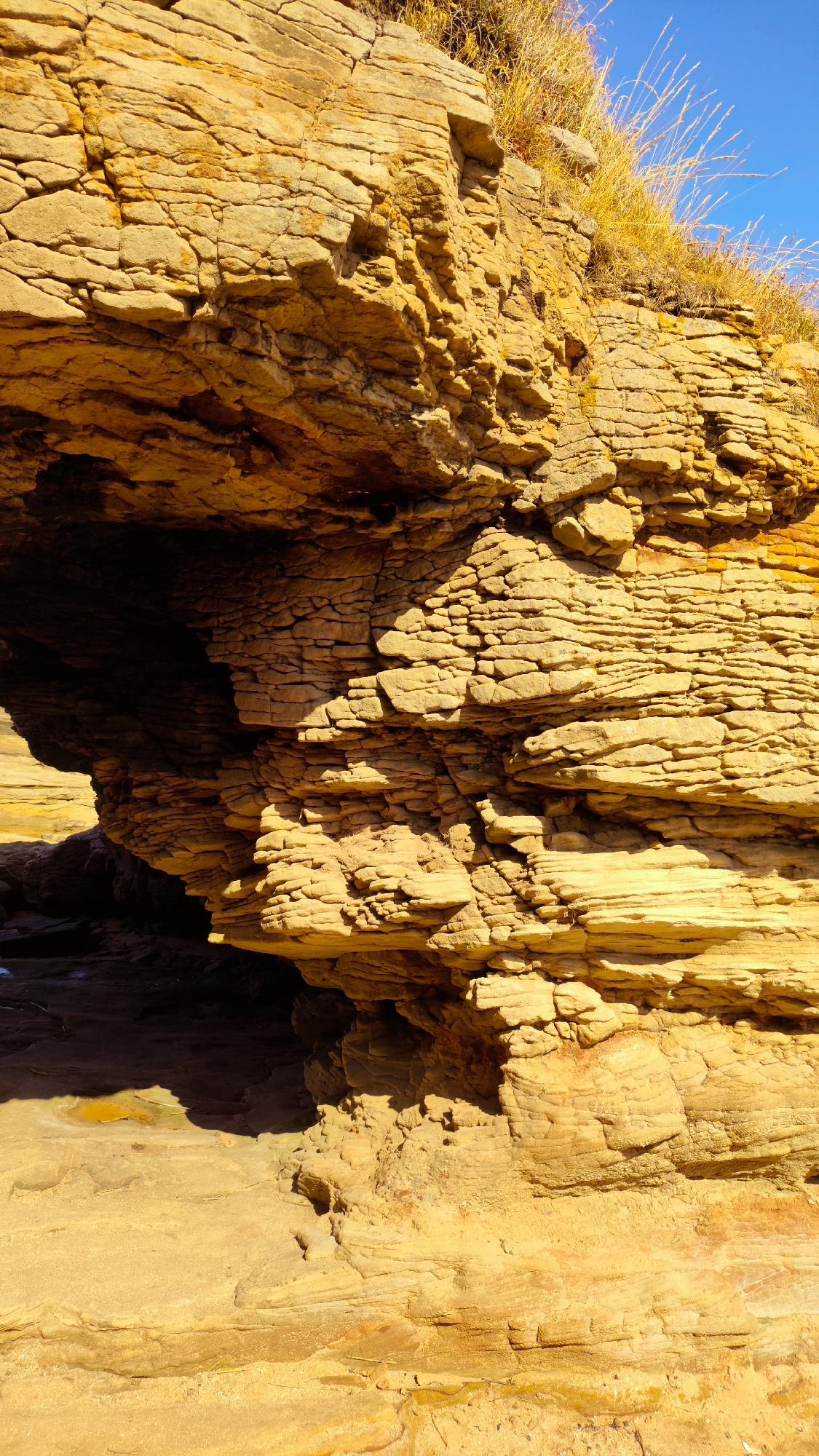 The image shows a close-up of layered, weathered sandstone formations at Red Rocks, West Kirby, United Kingdom. The rock displays a rough texture with horizontal striations indicating its geological history. Sunlight illuminates the right side of the rock face, casting shadows into the crevices and an overhang on the left. Dry, straw-like vegetation grows along the top edge of the rock formation. The ground in the foreground is sandy and light-colored, with some darker patches suggesting dampness or shadows. The sky above is a clear, bright blue.