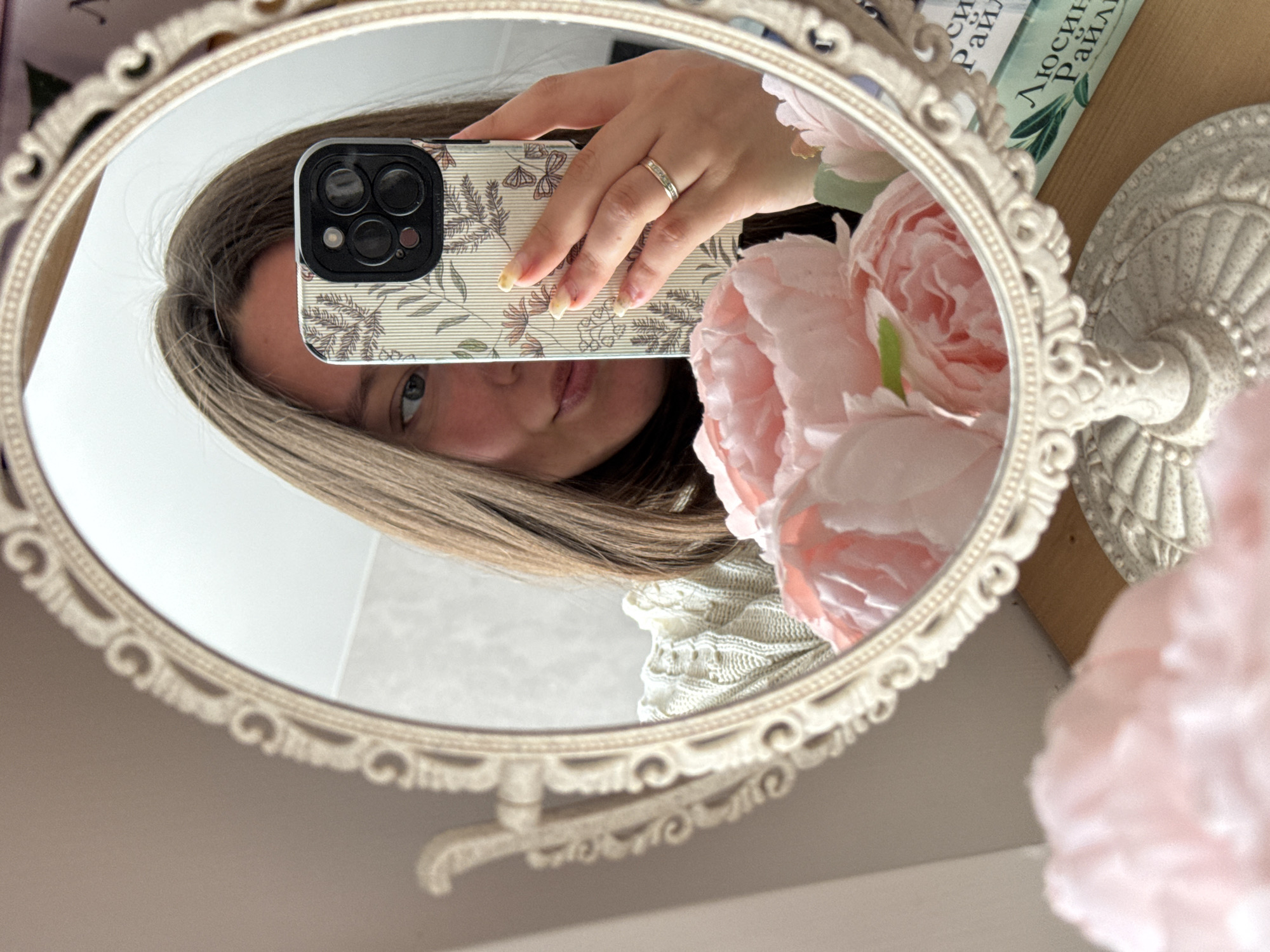 A close-up image captures a person taking a mirror selfie indoors. The central element is an oval-shaped mirror with an ornate, light cream-colored frame, reflecting the upper part of a person and her hand holding a smartphone.
The person, likely female, has long, light brown hair framing her face. Her right eye, nose, and lips are visible in the reflection, with a neutral and calm expression. She wears a textured, light-colored knitted garment. Her right hand, adorned with a simple silver-colored ring on her ring finger, holds a smartphone. The phone's multi-lens camera system is visible, and its case features a delicate botanical pattern with green foliage, pink flowers, and subtle butterfly motifs on a light background. Her fingernails are painted with a light color and appear somewhat grown out.
Beside and partially reflected in the mirror are several light pink artificial flowers, resembling peonies or roses, contributing a soft, decorative touch. To the right of the mirror, a light green book with white Cyrillic text is partially visible. The text clearly shows "ЛЮСИН РАЙЛ" (LYUSIN RAYL), indicating an author's name or part of a title. An ornate, light-colored decorative stand is also present on the right. The background behind the mirror is a plain light wall, and a wooden surface is visible beneath the book. The scene is brightly lit, suggesting daytime.