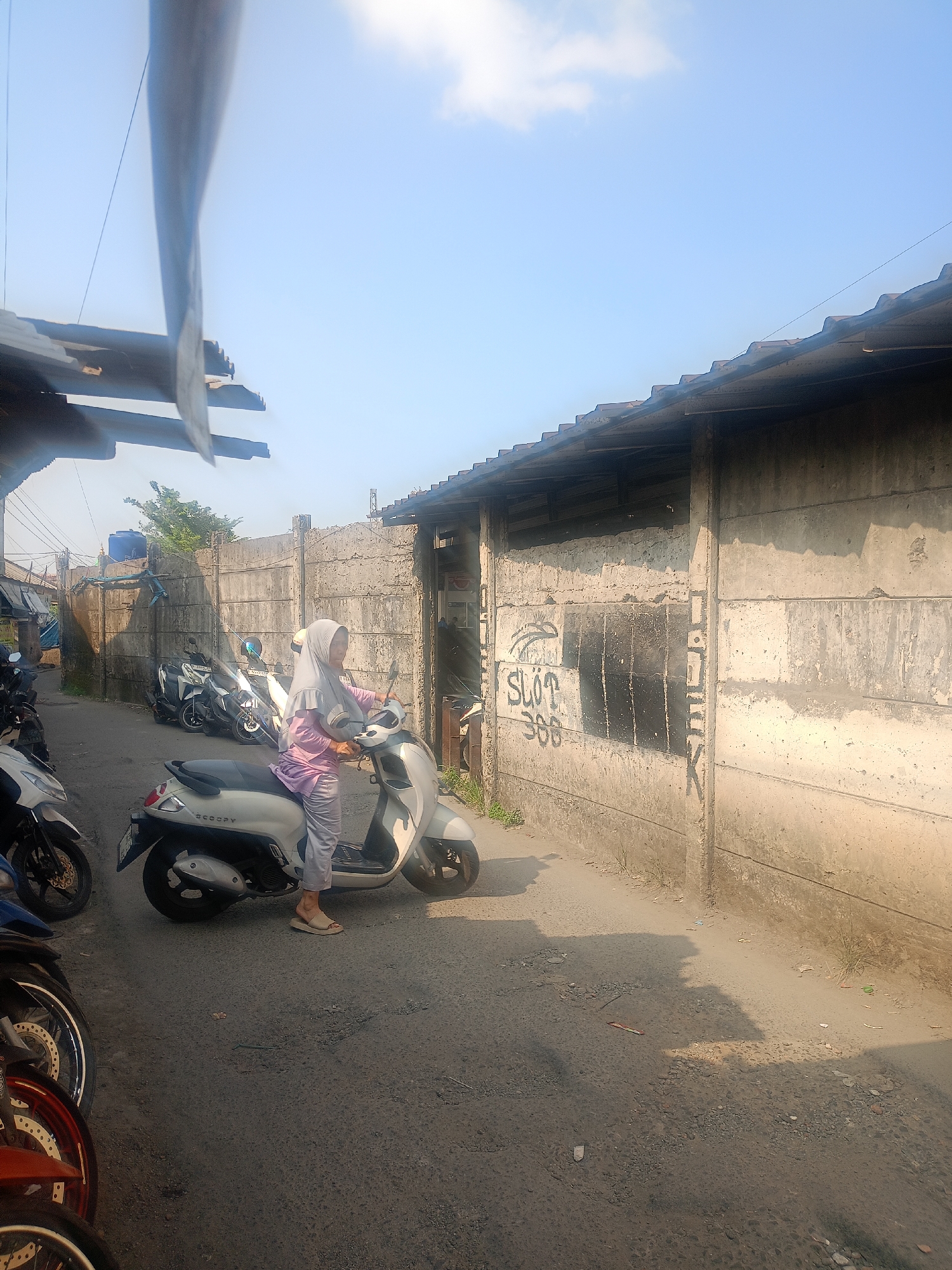 In Bojong Gede, Indonesia, on a sunlit day, a woman wearing a light purple headscarf and matching outfit is seated on a white scooter. She is positioned on the right side of a narrow, paved road, facing left. The road is lined with a tall, grey concrete wall on the right, which appears to be part of a building's exterior and has some graffiti, including "SLOT 300," and black spray-painted sections. The wall has horizontal seams and appears weathered. To the left of the road, several other motorcycles are parked, some partially visible. Beyond the wall on the right, a portion of a building with a corrugated metal roof is visible. The sky above is clear and blue with a few wispy clouds. The overall scene depicts a residential or commercial alleyway in Bojong Gede.