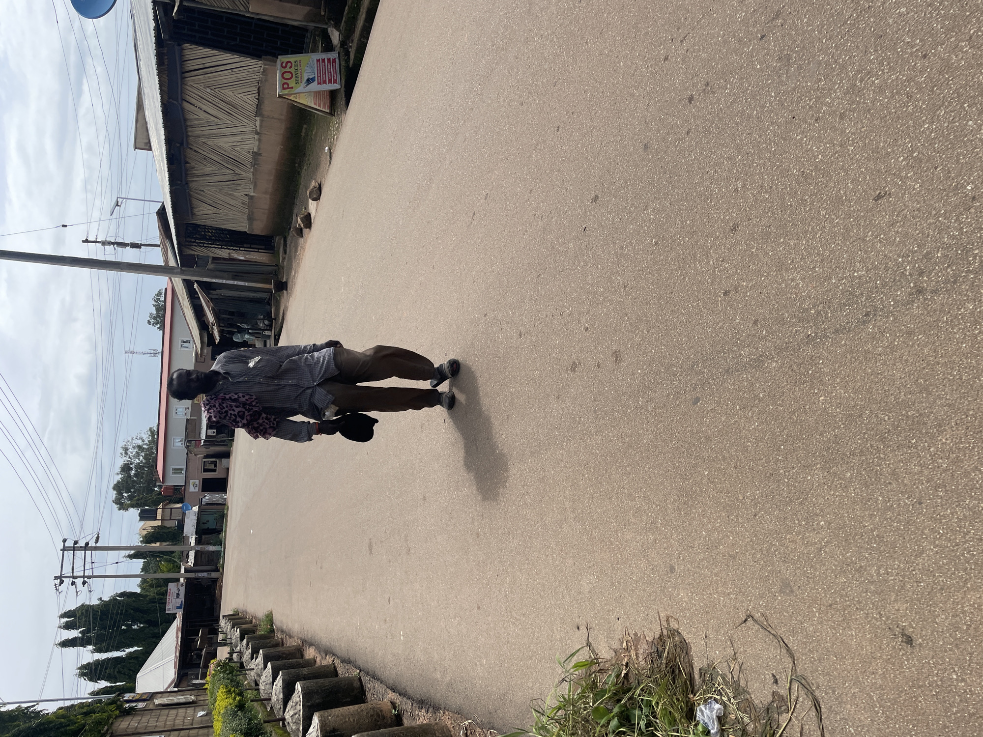 A man walks down a paved road in Jos, Nigeria. He is wearing a checkered shirt, brown pants, and sandals. He carries a dark hat and a patterned cloth draped over his left shoulder. The road is lined with short, cylindrical concrete posts on the left and buildings with corrugated metal roofs on the right. A sign for "POS SERVICES" is visible on the right side of the road. The sky is cloudy.