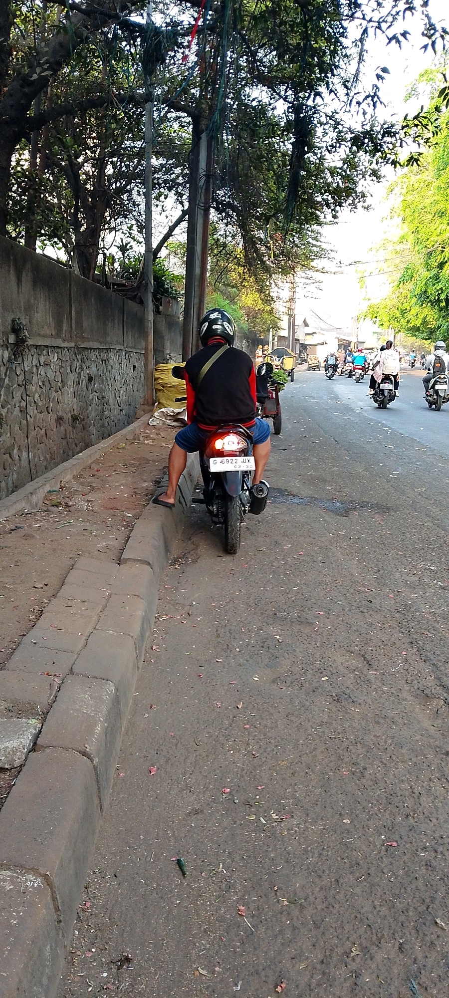 The image shows a street scene in Bekasi, Indonesia. In the foreground, a person is riding a black motorcycle, wearing a black and red jacket and a black helmet. The motorcycle has a license plate that reads "G 6922 JUK" and a sticker that says "03-20". The motorcycle is parked on the right side of a road, next to a concrete curb. The road is paved with asphalt and has some debris scattered on it. In the background, there are trees with green foliage, a stone wall, and other vehicles and pedestrians. The overall impression is that of a busy street in a tropical urban environment.