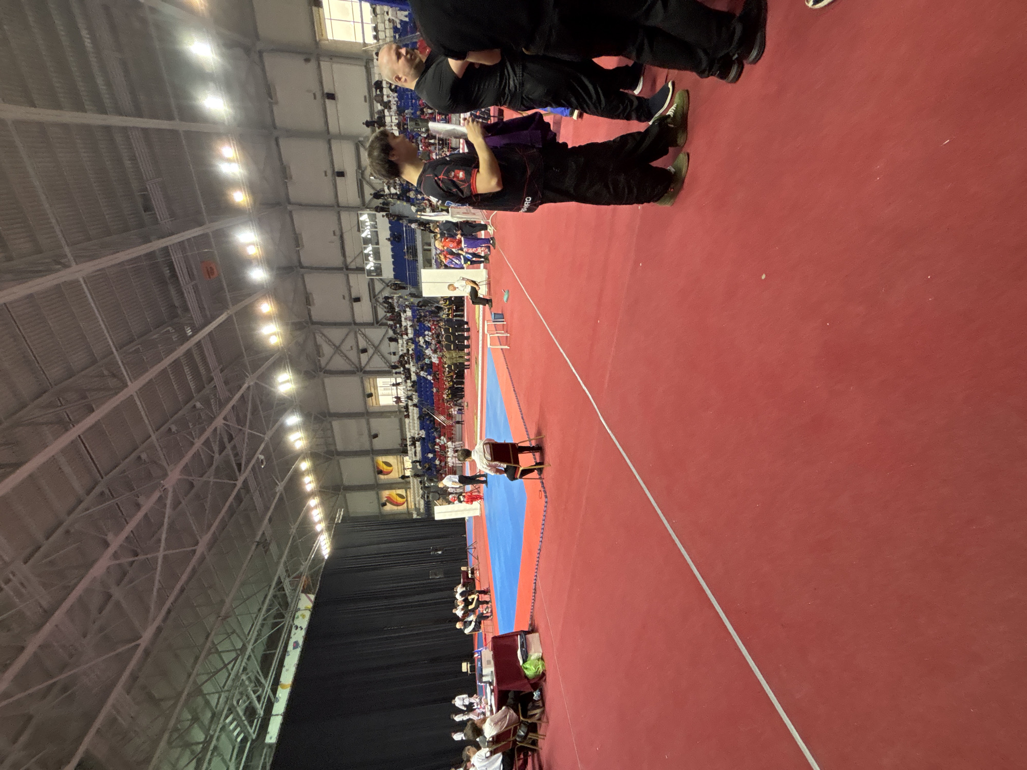 The image shows a large indoor arena, likely a sports venue. The floor is primarily red, with a blue section marking a competition area. People are present, both standing near the camera and seated in bleachers further away. Some individuals near the camera are wearing sportswear. The arena has a high ceiling with metal support structures and lighting fixtures. The location is the Perm Krai Sports Complex named after V.P. Sukhariev in Perm, Russia, as indicated by the signage.