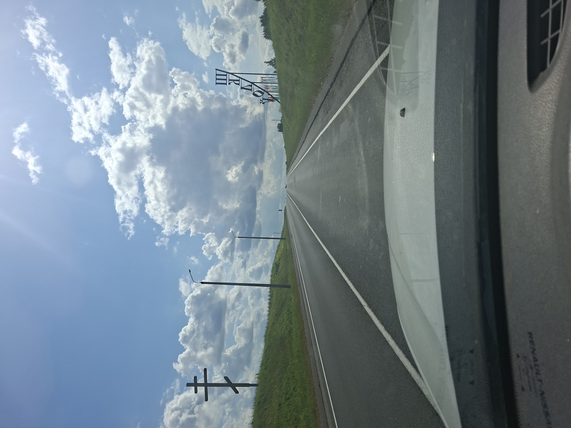 The image is taken from the perspective of someone inside a vehicle looking out through the windshield. The vehicle is driving on a paved road that curves slightly to the right. The road has two lanes in the direction of travel and is marked with white dashed lines. On the left side of the road, there is a grassy embankment with a large, dark-colored Orthodox cross. Further along the road on the left, there is a street lamp. On the right side of the road, the grassy embankment is topped with a large, abstract metal sculpture that appears to be made of vertical and diagonal bars, with some colorful flags at its base. The sky is blue with scattered white cumulus clouds, and the sun is shining brightly, creating a lens flare in the upper right corner of the image. The overall impression is of a sunny day on a highway in a rural or semi-rural area, with prominent religious and artistic landmarks visible. The location is identified as Starashklovski Selsoviet, Belarus, suggesting a rural setting.