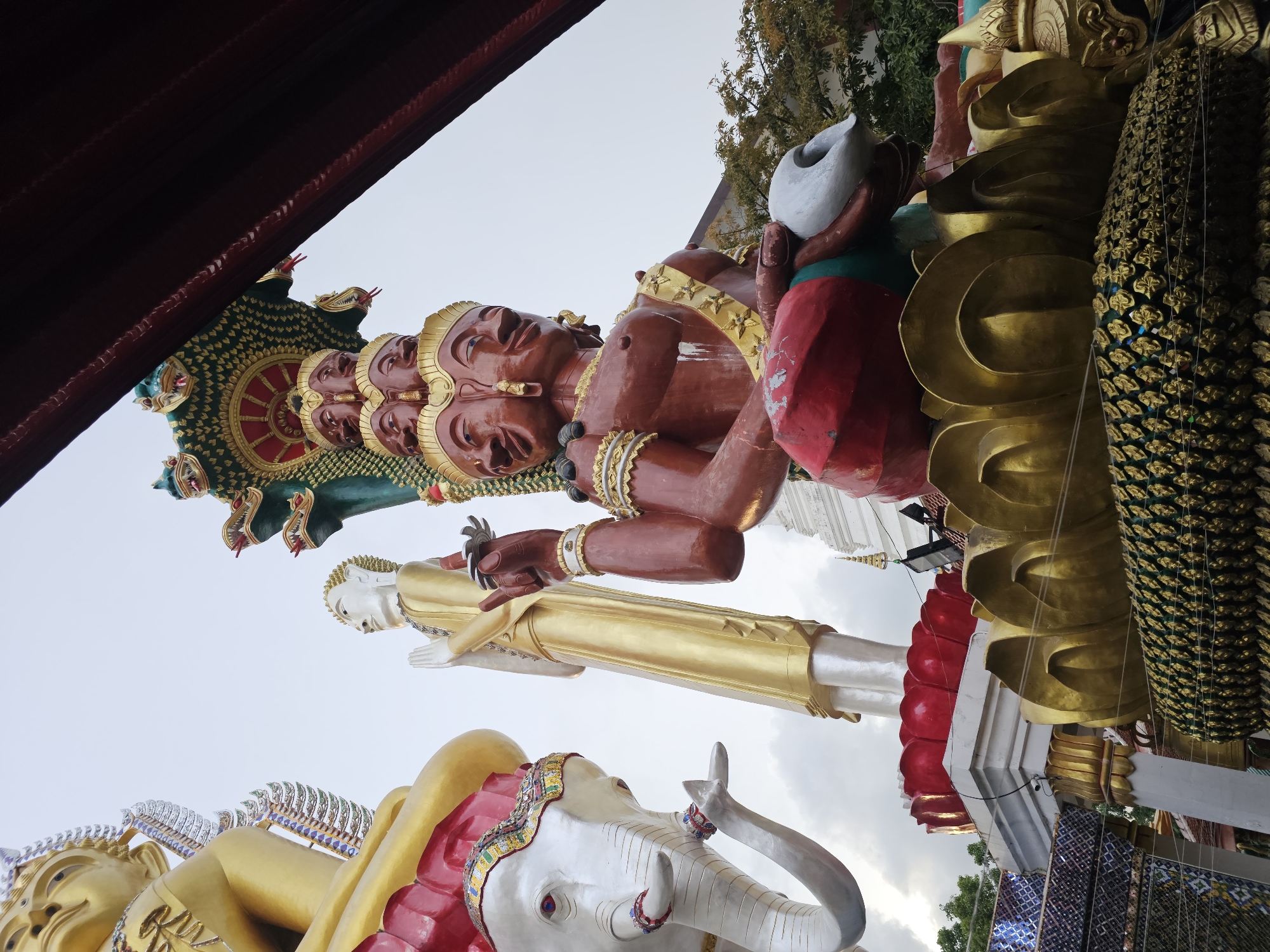The image depicts a low-angle view of several large, ornate statues in what appears to be an outdoor temple complex, likely in Bangkok, Thailand. In the foreground and to the right, a large, multi-headed, reddish-brown statue is prominent. This statue has multiple faces, and above it, a green multi-headed serpent or Naga creature is visible. The statue is seated on a golden lotus-like base. To the left, a white elephant statue with red accents and decorative elements is positioned. Behind the elephant and slightly to the right of it, a tall, golden statue of a figure in prayer is seen. Further in the background, glimpses of white temple structures and trees are visible. The sky is overcast. The scene is rich in religious iconography, typical of Thai Buddhist temples.