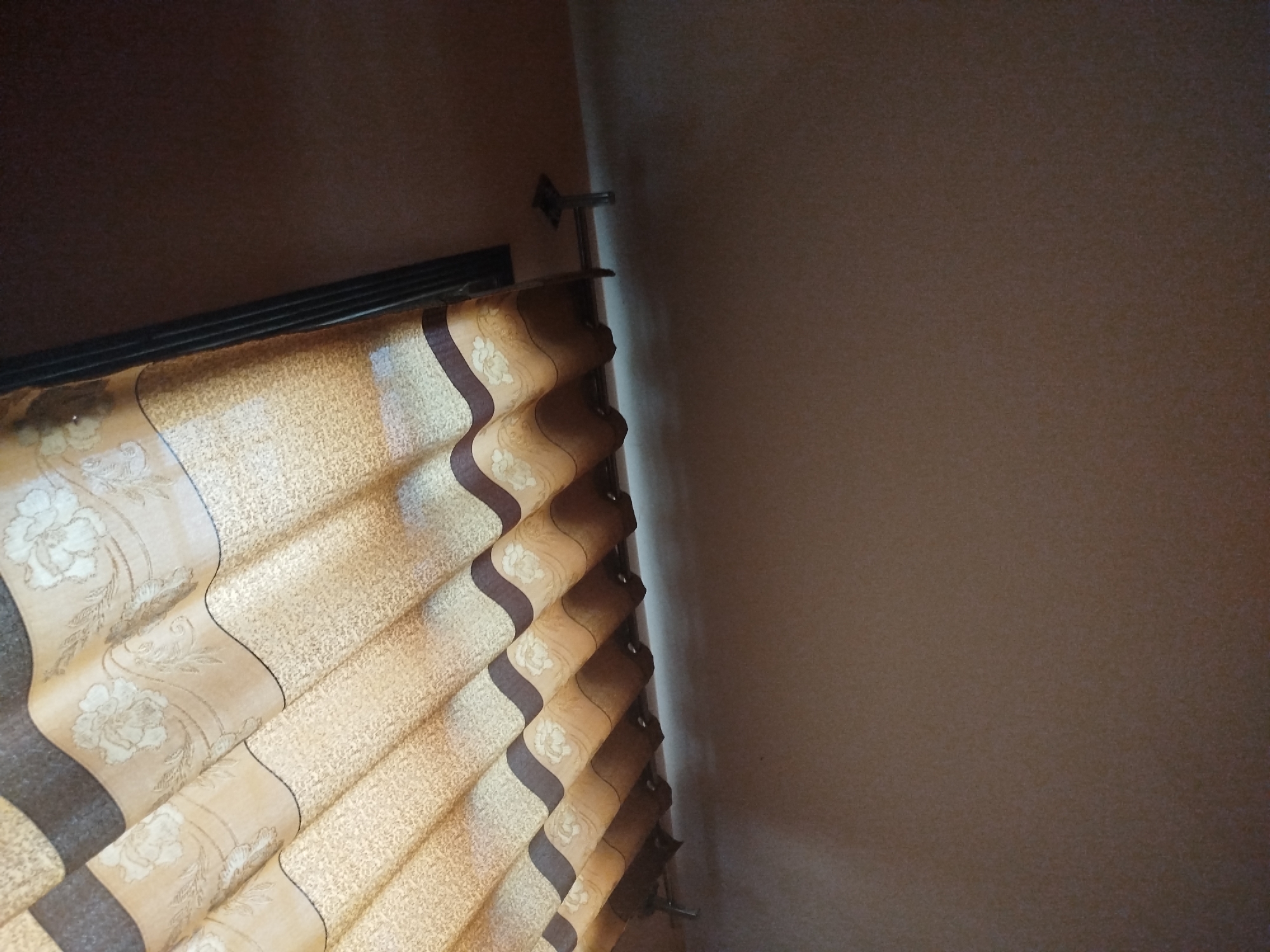 This is a close-up, low-angle shot of a window with closed blinds and a dark brown wall. The blinds are made of a light beige fabric with a textured weave and a decorative pattern of pale flowers and thin dark brown stripes that create a zig-zag design. The blinds are pleated and hang from a dark metal rod supported by brackets attached to the wall. The wall to the right of the blinds is a solid, muted brown color. The top left corner of the image shows a dark brown ceiling. The lighting suggests an indoor setting, possibly during the day given the light filtering through the blinds, though no direct light source is visible. There are no people or animals present. The scene is static and appears to be a corner of a room. No text is visible in the image. The location context provided is Lassan, Pakistan.
