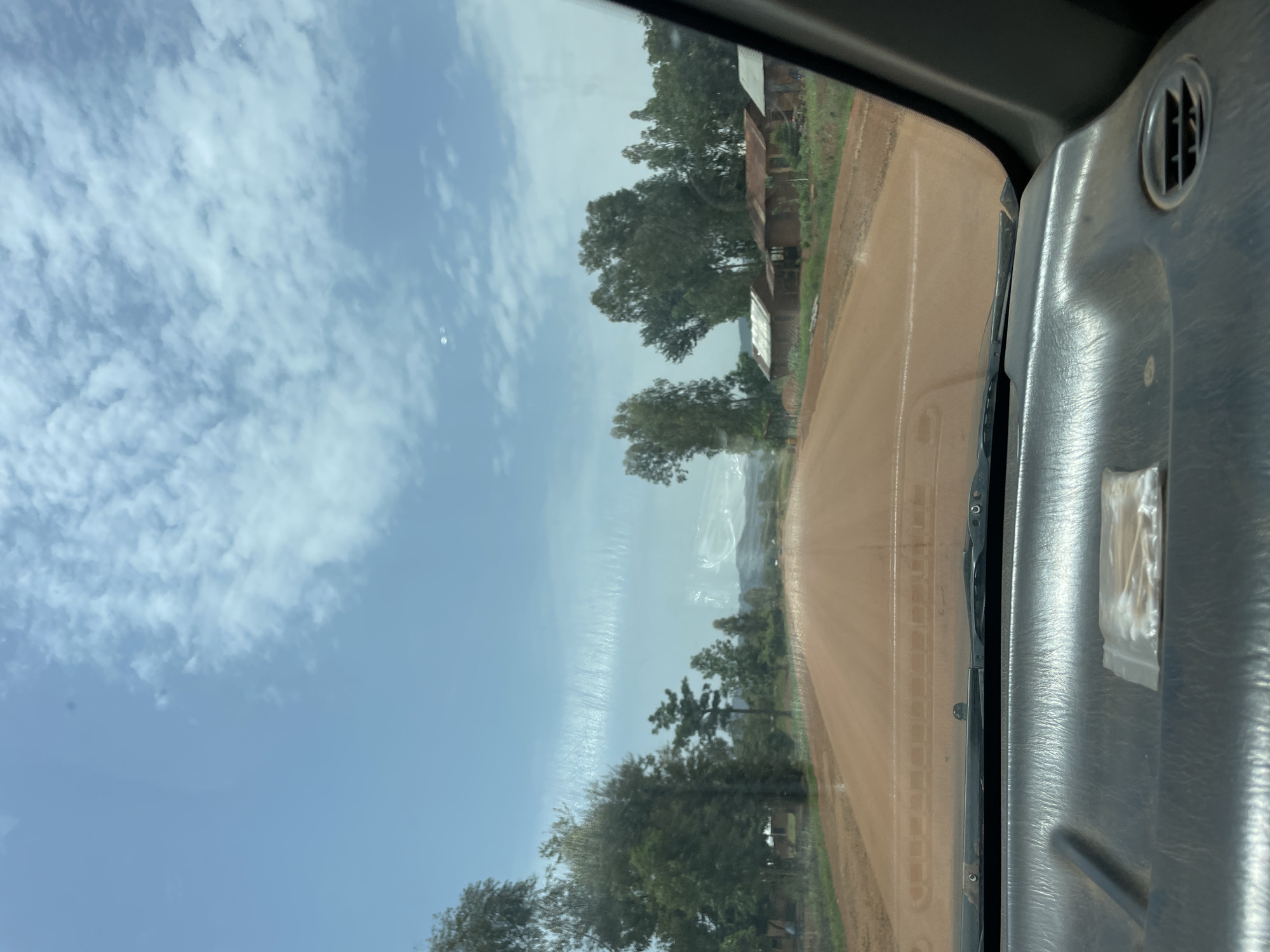 The image, taken from inside a vehicle, depicts a dirt road in a rural environment. On the left, there is a blue sky with scattered white clouds. Trees and sparse vegetation line the side of the road. Some structures are visible amongst the trees. The dashboard of the vehicle is visible on the right side of the frame. The windshield has a visible water stain. It is not possible to confirm this image was taken in the city of Zankan, Nigeria, or to discern people, specific actions, or interactions within the scene.