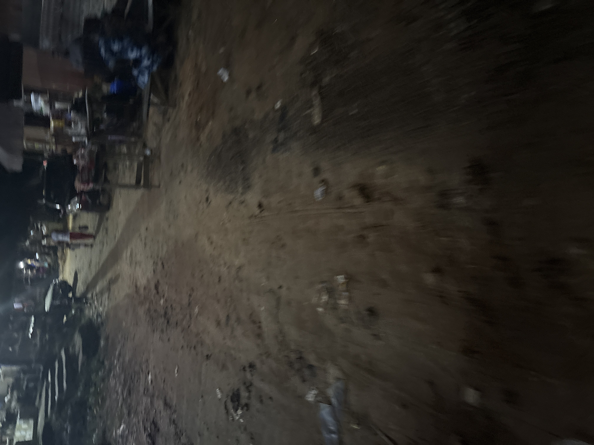 The image depicts a dimly lit, outdoor scene likely in the city of Onitsha, Nigeria, suggesting a marketplace or a busy street at night. The ground is unpaved and appears to be dirt or mud, with scattered debris including what looks like plastic bags and other refuse.
In the background, several structures are visible. To the left, there are steps leading up to buildings. Further back and to the right, there are market stalls with tables laden with various items, some appearing to be food or goods for sale. A person is standing near one of the stalls, and another figure, possibly a child, is walking away from the camera. A vehicle, possibly a car or a van, is parked on the street, and a motorcycle is also visible. Artificial lighting from lamps and possibly streetlights illuminates the scene unevenly, casting long shadows and creating a somewhat blurry effect due to the low light conditions and possibly motion. The overall impression is one of a vibrant, albeit somewhat disorganized, nocturnal commercial activity.