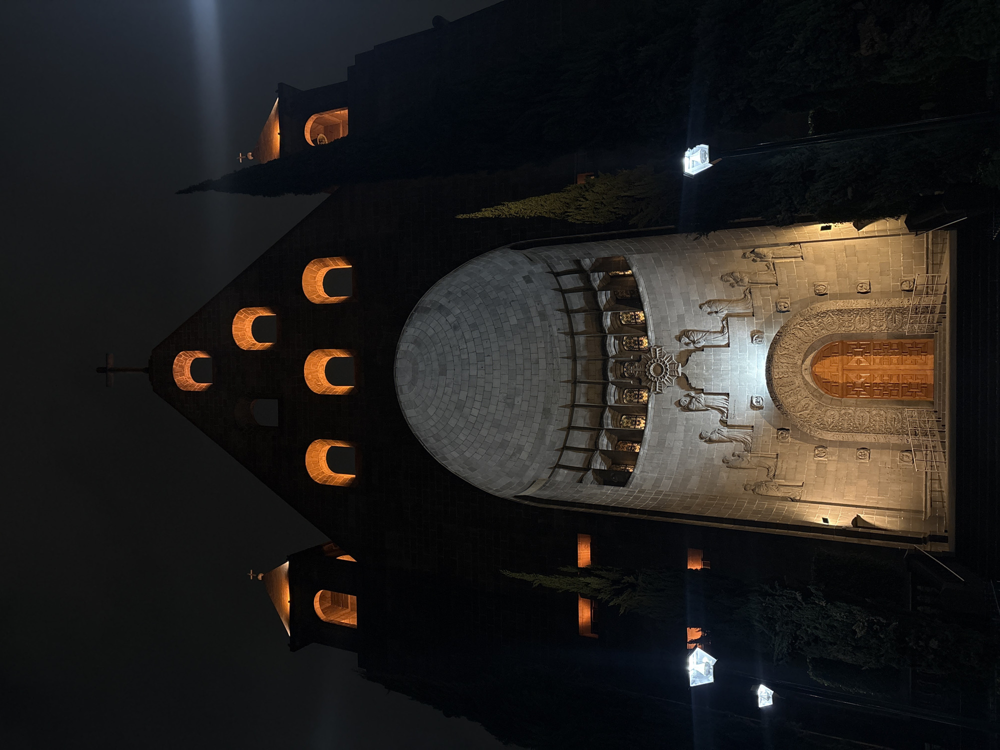 Here is a description of the image:
The image shows the Monument to the Niños Héroes in Mexico City at night. The monument is illuminated with lights, highlighting the stone structure and decorative carvings. The architecture is a mix of angular walls with semi-circular windows and a rounded facade. The scene is taken from a high angle, showing the monument's layout and surrounding trees. There are several light posts near the monument.