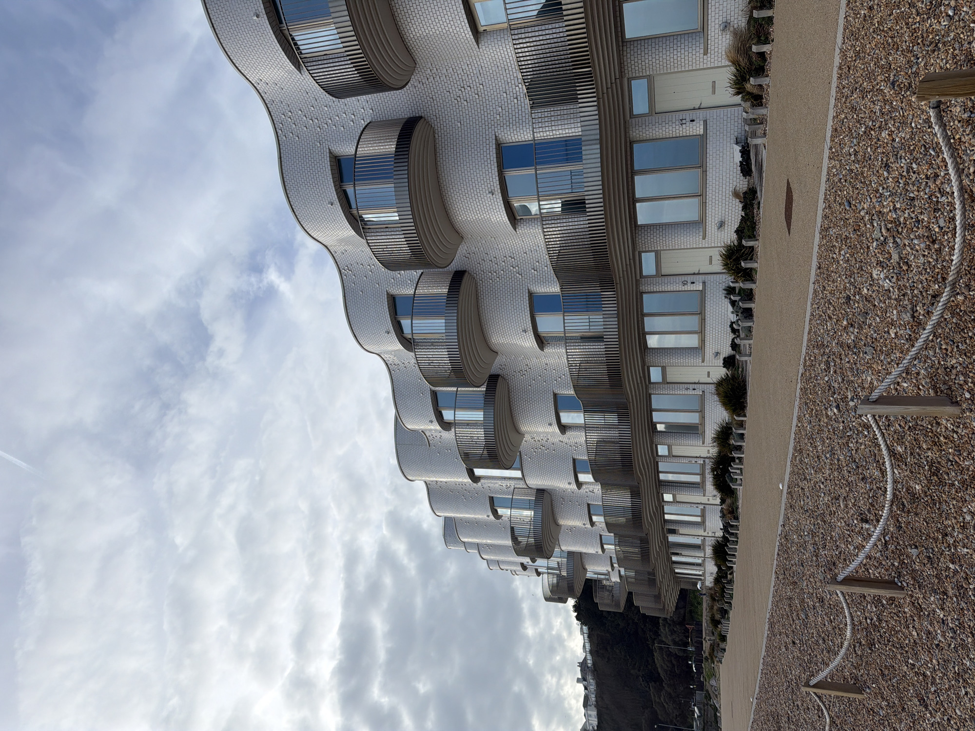The image shows a row of modern buildings along a beach in Folkestone and Hythe District, United Kingdom. The buildings feature white brick exteriors, distinct curved facades, and large windows reflecting the sky. A paved walkway runs along the base of the buildings, separating them from a pebble beach with wooden posts and ropes. The sky is overcast with clouds.