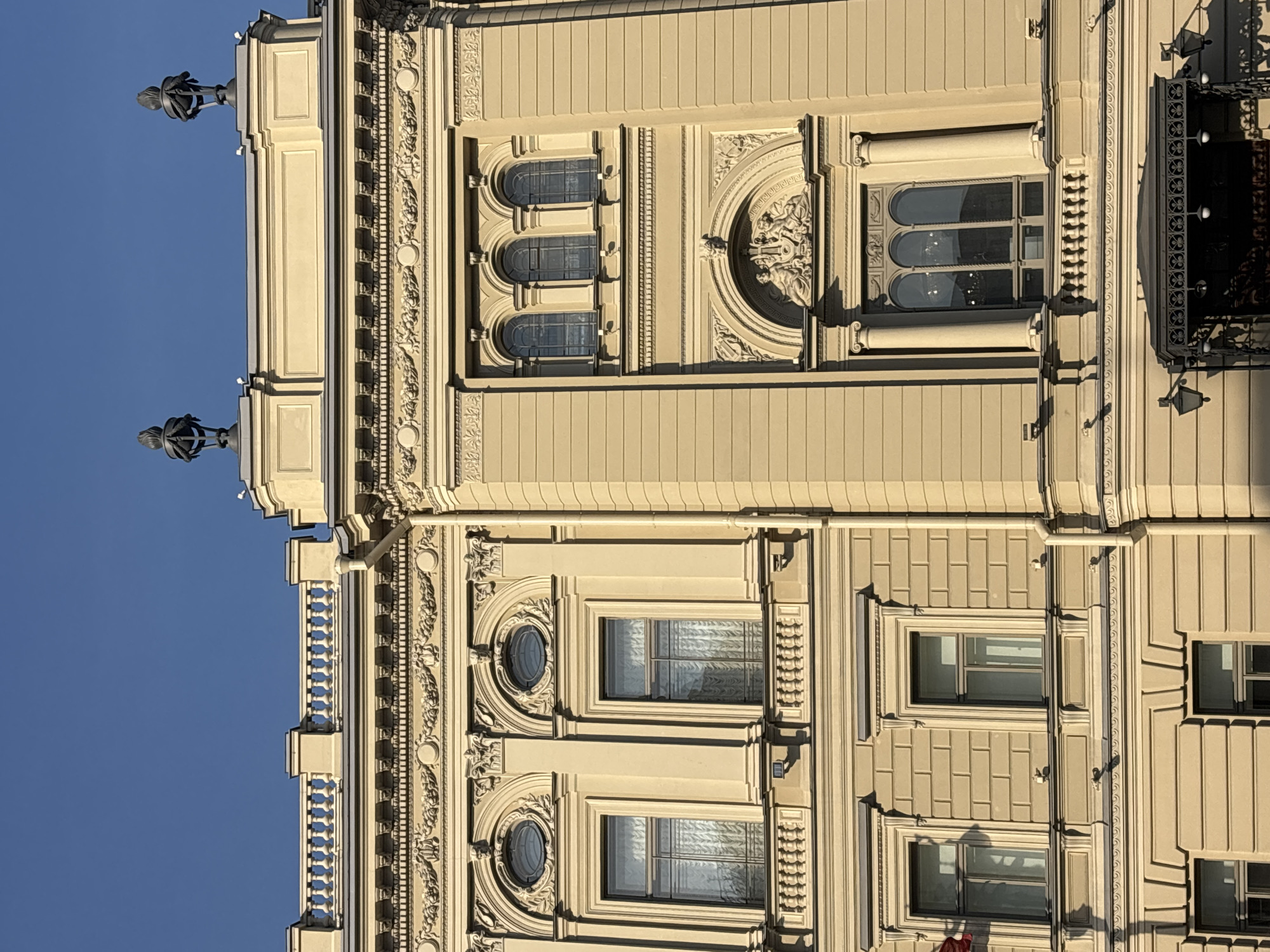 The image shows the facade of a grand building, likely an opera house or theater, in Saint Petersburg, Russia. The architecture is neoclassical, characterized by its ornate detailing, pilasters, arched windows, and balustrades. The building is bathed in sunlight, casting shadows and highlighting the architectural features. The sky is clear blue.
Specifically, the building appears to be the Mariinsky Theatre, a renowned opera and ballet theatre located at Theatre Square in Saint Petersburg. The facade displays classical architectural elements such as decorative friezes, cornices, and sculpted pediments. Several windows are visible, some with arched tops and intricate tracery. On the rooftop, decorative urns or finials can be observed. The lower portion of the building includes a covered entrance with ornate metalwork and hanging lamps, suggesting a formal entryway. The overall impression is one of opulence and historical significance. There are no people or active events depicted in the image, focusing solely on the architectural details of the building.