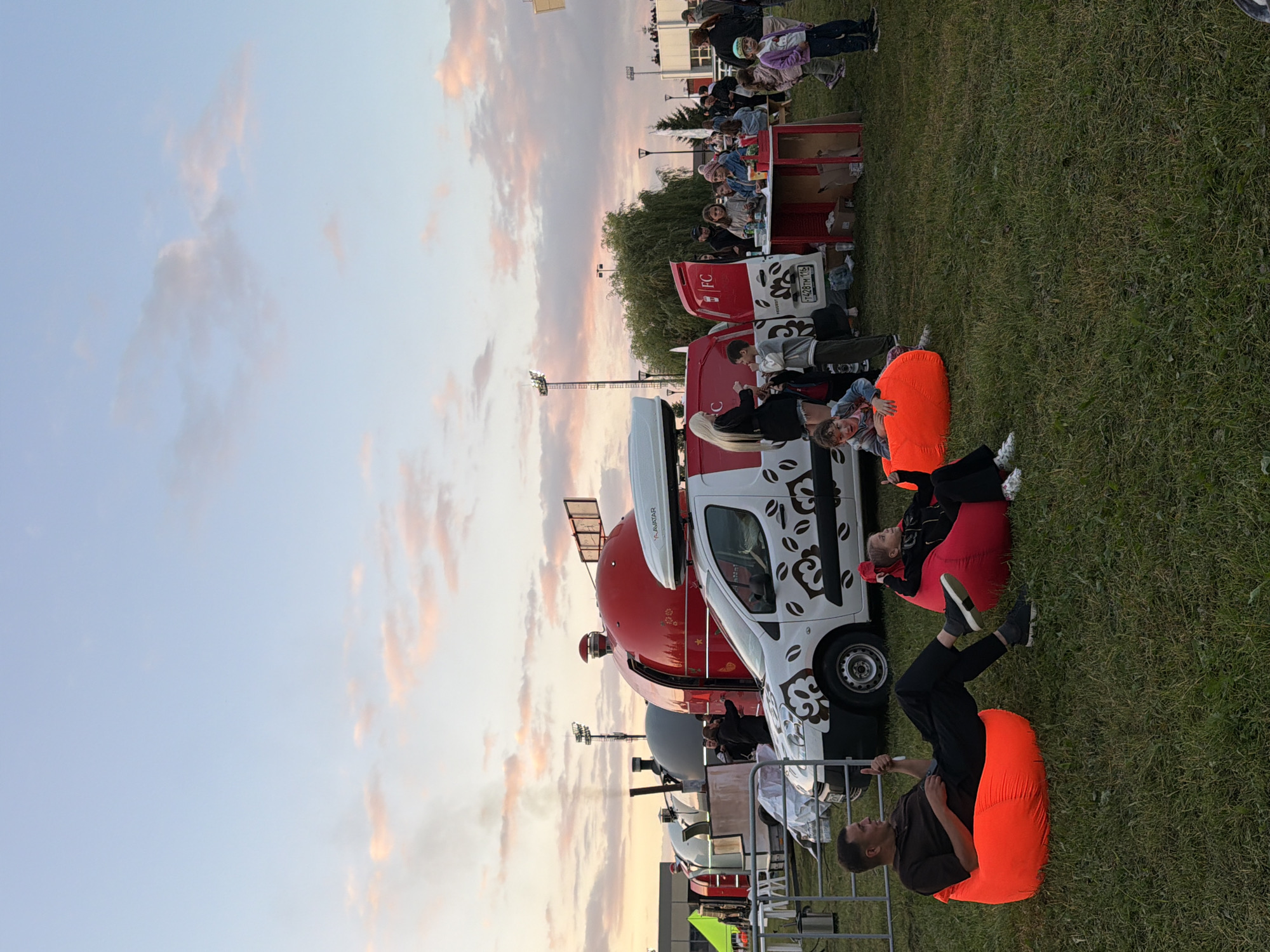 In an outdoor grassy area, a diverse group of people are gathered near several food trucks under a pale blue sky with wispy pink and orange clouds, indicating dusk. In the foreground, a man is seated on an orange beanbag chair, giving a thumbs-up, while a young girl in a pink beanbag chair nearby looks towards the camera. Behind them, a white van with a coffee bean decal is parked, its side door open, revealing people interacting. Further back, various food trucks with distinctive dome-shaped roofs are visible, along with a group of children and adults congregating around a red table setup. The overall scene suggests a casual outdoor event or festival.
