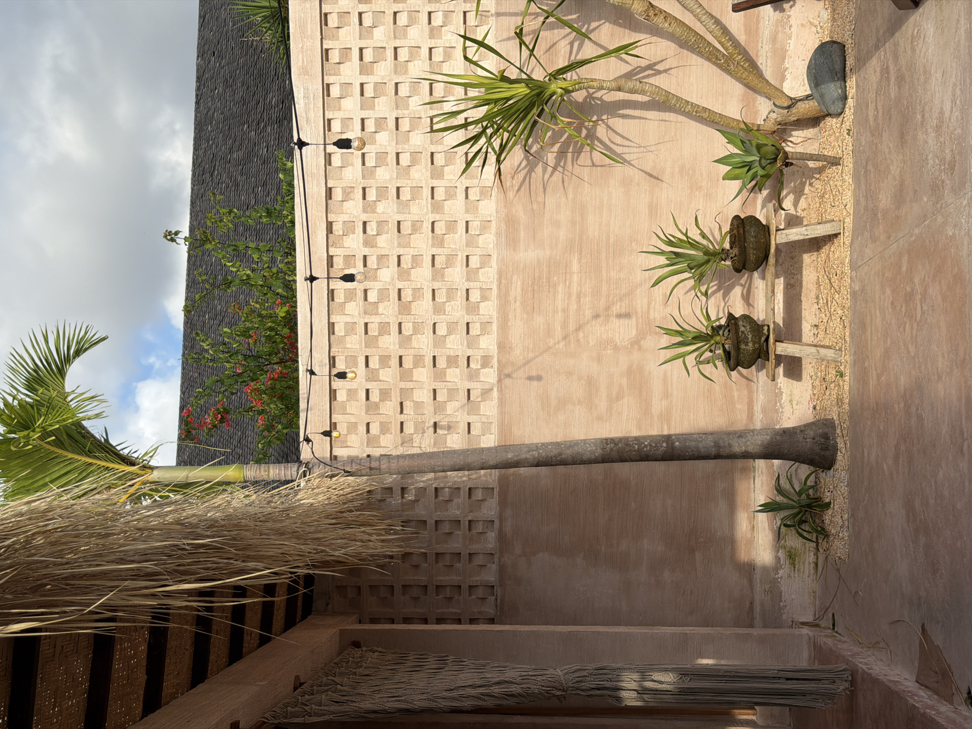The image, taken in North Kuta, Indonesia, shows a textured light brown wall composed of alternating square block panels and patterned panels. String lights run vertically down the wall, partially obscured by a leaning palm tree trunk. To the right of the wall, plants are arranged, some in pots and others directly on the ground. Below the wall, an area is covered by a thatched roof and a net hammock. A palm tree with green fronds is on the left. The sky is visible at the top left corner, with a mix of clouds and blue sky. A building with a dark grey roof is also visible in the background.