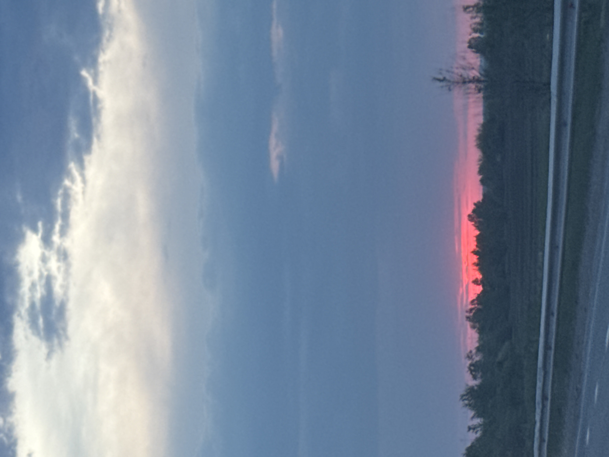 The image shows a landscape at sunset in Кондратово, Russia. A section of road is visible in the bottom right, with a guardrail running along the edge. Beyond the road, there is a line of trees silhouetted against the pink and red sky of the setting sun. The sky above is predominantly blue with patches of white and gray clouds.