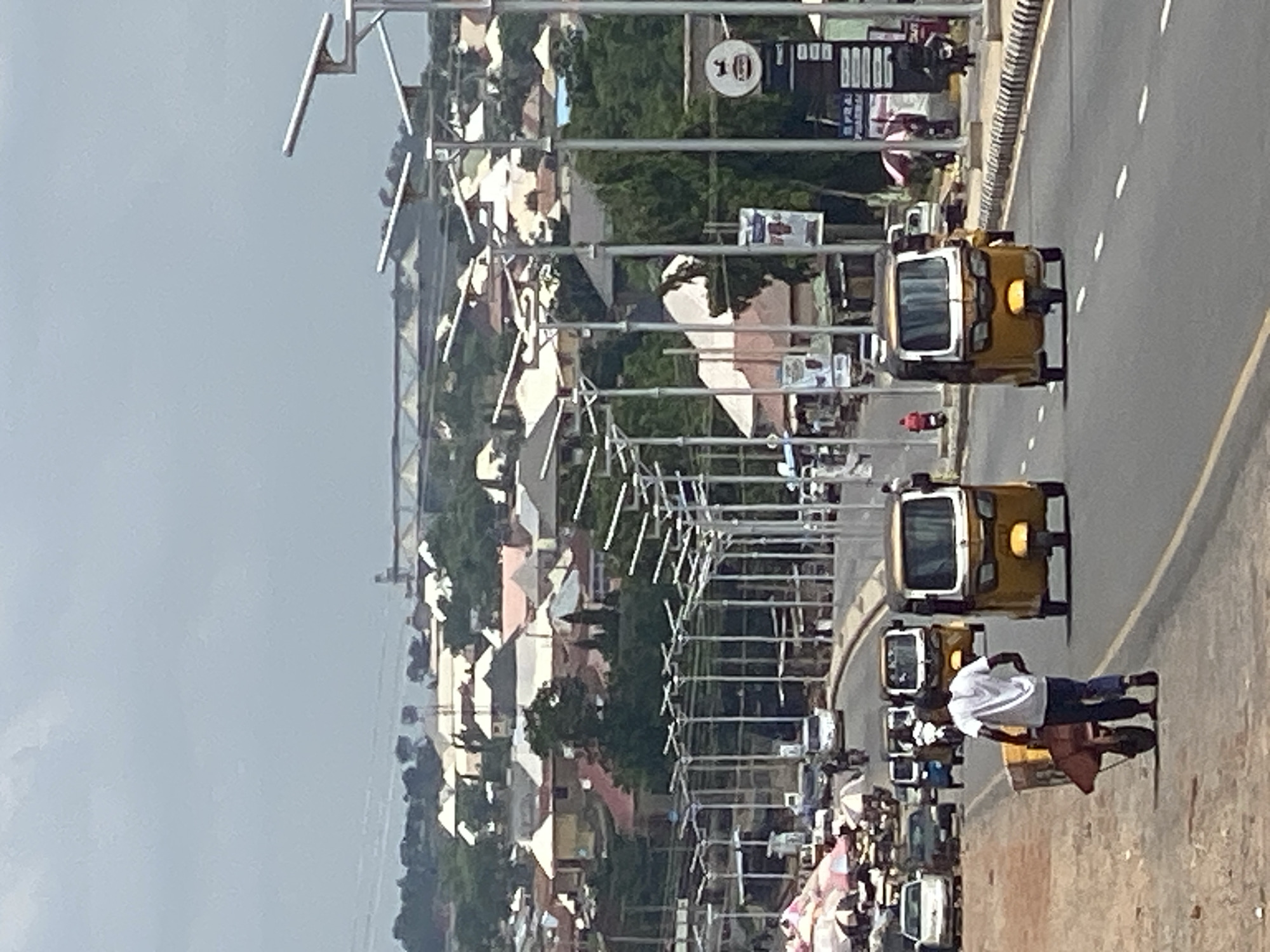 The image shows a street scene in Adankolo, Nigeria. The main focus is on the yellow "keke" tricycles that are a common form of transportation. A man wearing a white shirt and dark pants walks along the side of the road, pulling a brown cart. Numerous keke tricycles are on the road, some moving and some seemingly stopped. A town is in the background with light-colored buildings and trees. A series of metal posts and crossbars runs parallel to the road. There is a speed limit sign visible.