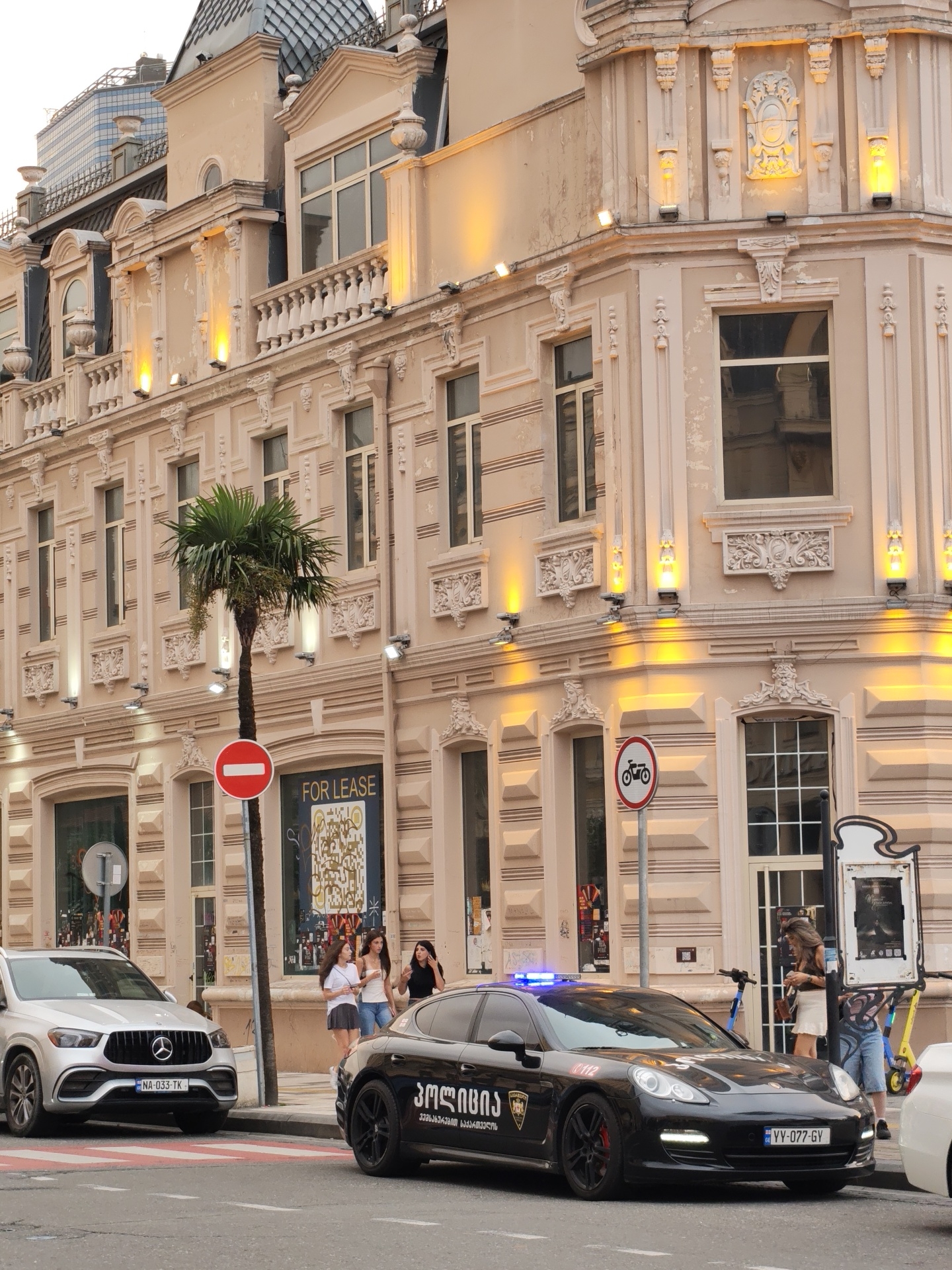 In Batumi, Georgia, on a city street, a black Porsche Panamera police car with flashing blue lights is parked on the right side of the frame. The car has Georgian script on its side and a license plate that reads "YY-077-GV." A white Mercedes SUV is parked on the left, with a license plate "NA-033-TK." Three young women stand on the sidewalk near a palm tree and a "FOR LEASE" sign. A man stands near a bicycle sign and a poster display on the right side of the street. Several electric scooters are parked near the man. The background features a light pink, ornate building with windows, balconies, and decorative elements, illuminated by yellow lights. The sky is bright, suggesting daylight.