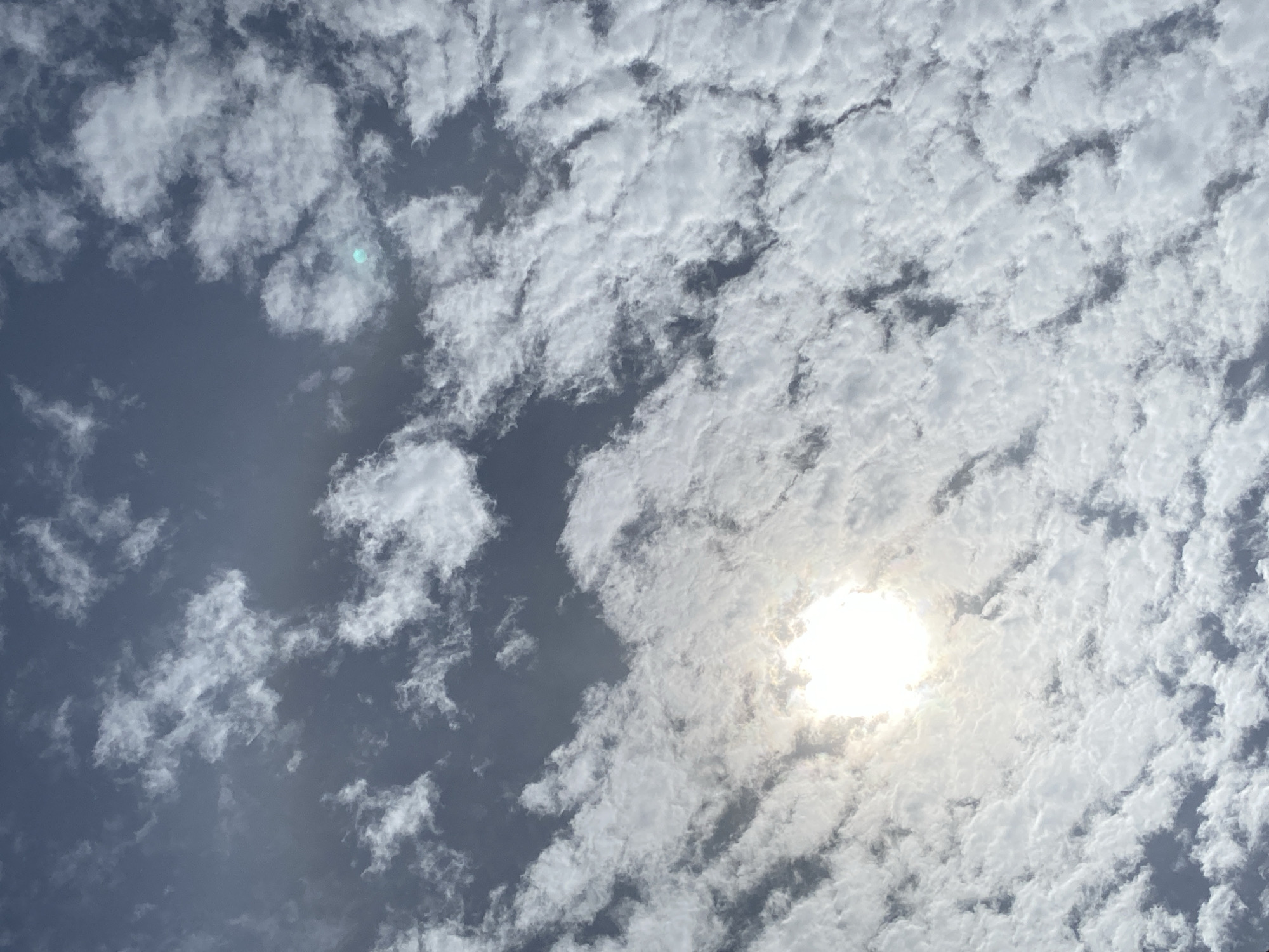 The image is an outdoor, low-angle shot of a sky filled with clouds and the sun. The sun is a bright, concentrated light source in the lower left quadrant of the image, emitting rays and creating a halo effect. Numerous white clouds, some dense and textured, others wispy, fill the frame, interspersed with patches of darker, grey sky. One small, bluish lens flare is visible in the upper right quadrant. The scene appears to be captured during daytime. The location is listed as Unknown, Unknown.