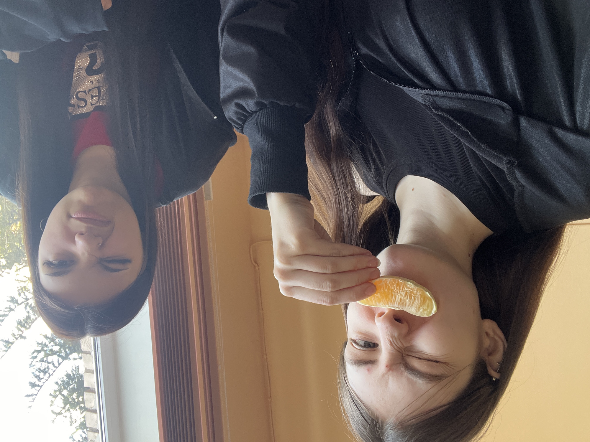 The original image is rotated 90 degrees clockwise. When viewed upright, it features two young women indoors in Saint Petersburg, Russia, captured from a slightly low angle looking upwards.
On the right, one woman with long dark hair is playfully holding an orange segment over her right eye, squinting her face and winking with her left eye to create a humorous expression. She is wearing a dark, long-sleeved top with a zippered detail on the shoulder.
On the left, another woman with long dark hair smiles slightly at the camera, winking with her right eye. She is dressed in a dark, long-sleeved top with a red layer visible underneath. The word "GUESS," embellished with sparkling rhinestones, is clearly visible on her chest (appearing upside down in the original rotated image).
The background on the left features a window revealing bare tree branches against a bright sky, indicative of daytime during a colder season like winter or late autumn. To the right of the window, a warm-toned wall or wooden paneling is visible. The overall mood is playful and lighthearted.