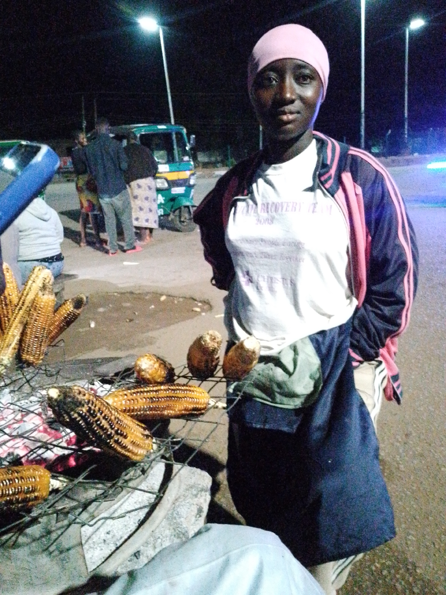 A young woman, wearing a pink head covering, a white t-shirt with visible text, and a dark jacket with pink stripes, stands at night. She appears to be a street vendor, with grilled corn on a wire rack in the foreground. The corn is golden brown and appears to be roasted over coals, some of which are visible underneath the rack. Behind her, a green and yellow tricycle, likely a commercial vehicle, is parked on a paved street. Several other people are visible in the background, some standing near the tricycle and others further away. The scene is illuminated by streetlights, creating a nighttime ambiance. The weather appears to be clear. The woman has a neutral expression. The text on her t-shirt is partially visible and reads "RECOVERY TEAM 2018". The setting suggests a market or roadside area in Bukuru, Nigeria.