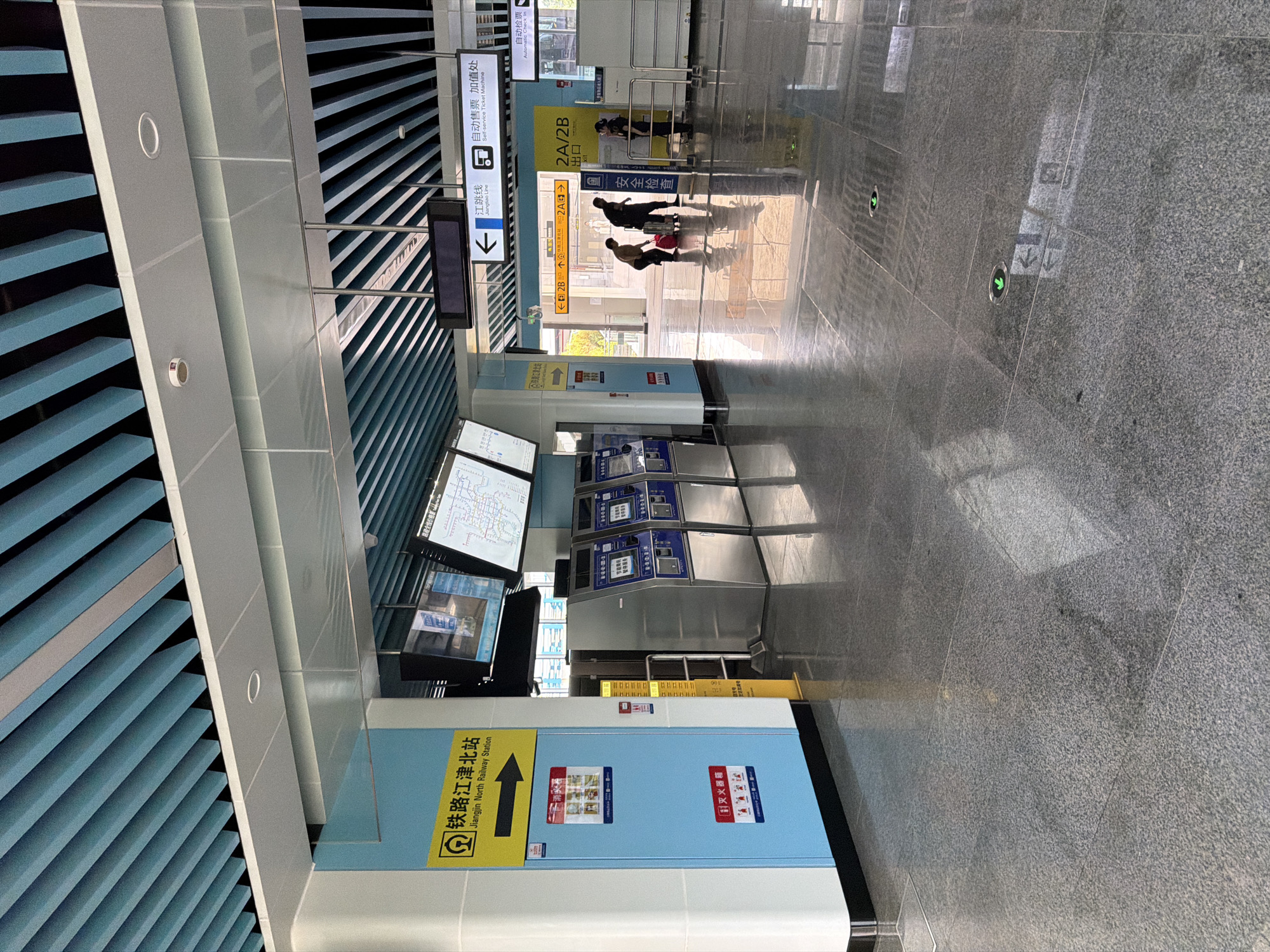 The image shows the interior of the Jiangjin North Railway Station in Jiangjin, China. The scene is brightly lit and features a modern, clean aesthetic with a polished grey stone floor. Above, a blue slatted ceiling directs the viewer's eye.
On the left side of the image, a light blue wall is adorned with yellow and red signage. A prominent yellow sign with a black arrow indicates the direction to "Jiangjin North Railway Station." Below this, a light blue door has multiple red and white signs with instructional graphics.
In the center of the image, a row of silver and blue self-service ticket machines is visible. Beyond the ticket machines, a set of automatic gates leads to an exit area.
In the background, several people are visible. Two individuals, one with a red suitcase, are walking towards the exit. Another person is standing near the automatic gates, seemingly interacting with a security guard. Signage in the background indicates "Jiangtao Line" and "Automatic Check In."
The overall impression is that of a functional and efficient public transportation hub.