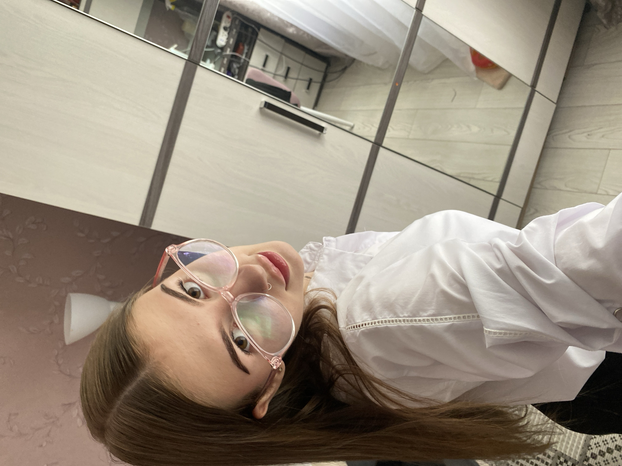 In a domestic indoor environment, possibly a bedroom in Dyakovo, Russia, a young woman with long brown hair is positioned facing the camera at a slightly upward angle. She is wearing a white collared shirt and pink-rimmed glasses. Her gaze is directed towards the lens, and she has a subtle smile. The background features a wardrobe with mirrored doors, reflecting the room. One mirror shows a glimpse of a dresser with drawers and some personal items. The woman's right shoulder and upper arm are visible, along with her hair falling over her left shoulder. The overall lighting is soft, suggesting it might be daytime.