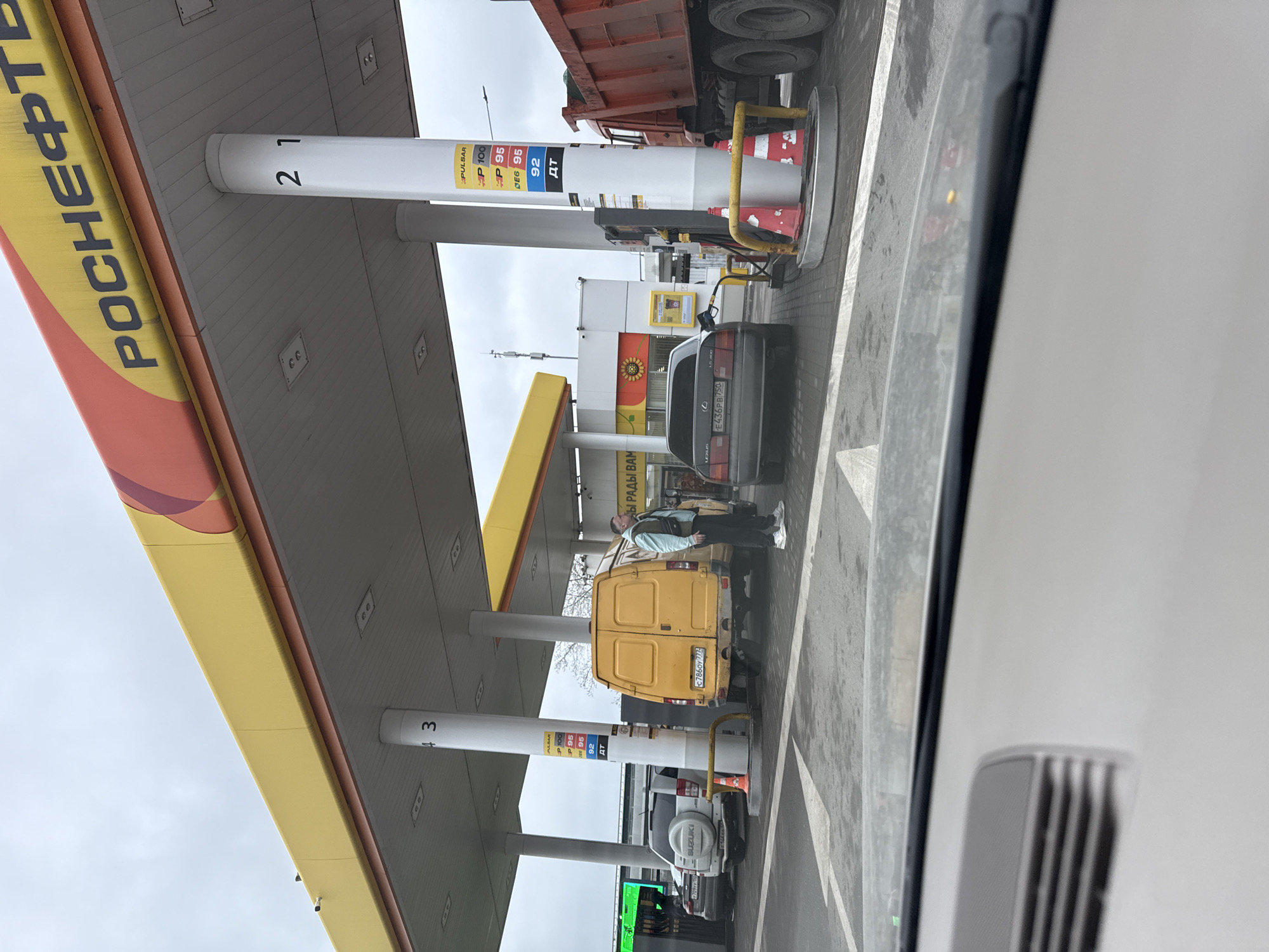 The image shows a gas station in Moscow, Russia, with a "Rosneft" logo. Station numbers 2 and 3 are visible on the support pillars. A person in a light blue jacket is standing next to a yellow van with the license plate "E786OY 777" at pump 3. A gray Lexus with the license plate "E436PB 750" is parked at pump 2. Behind the Lexus is a red truck. A white Suzuki SUV is parked behind the yellow van. Fuel prices are displayed on the pumps. The image was taken from inside a car.