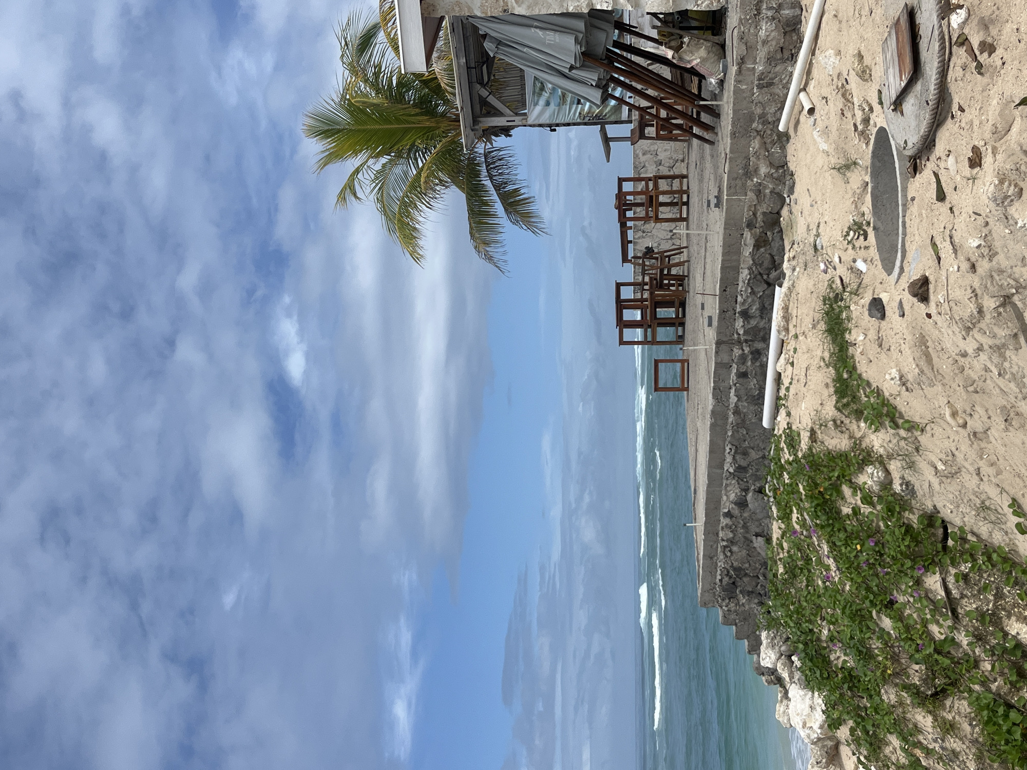 The image depicts a coastal scene likely situated in Kuta Selatan, Indonesia, characterized by a juxtaposition of natural and man-made elements. The foreground features a sandy area with trailing vegetation. Adjacent to this is a low wall or structure, likely a retaining wall. Further inland are several wooden chairs and tables, seemingly part of a beachfront establishment. A palm tree is partially visible, leaning toward the seating area. In the distance, the ocean meets the sky, with visible waves. The sky is partly cloudy.