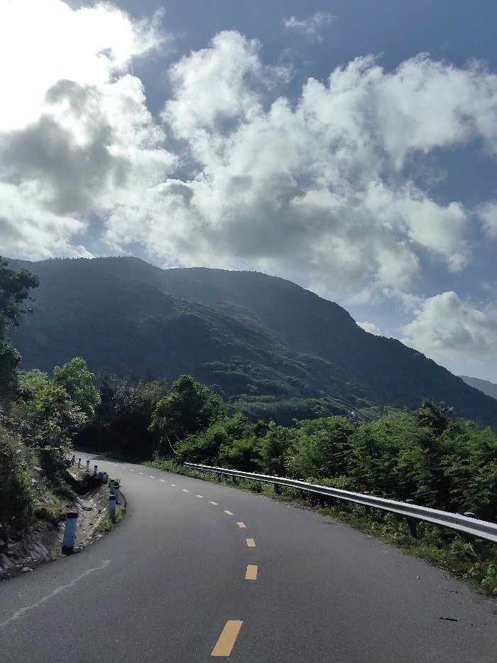 This image displays a winding asphalt road traversing a mountainous region in Ninh Hòa, Vietnam. The road, marked with a dashed yellow center line, curves sharply to the left in the foreground. Along the left edge of the road, a dirt embankment features several white concrete bollards with red tops and a visible drainage channel. The right side of the road is bordered by a silver metal guardrail, behind which dense green trees and vegetation are present. Large, dark green mountains, heavily covered in trees, dominate the background. The sky above is partly cloudy, showing patches of blue sky interspersed with white and grey cumulus clouds. No individuals, vehicles, or specific activities are observable. The scene is illuminated by natural daylight.