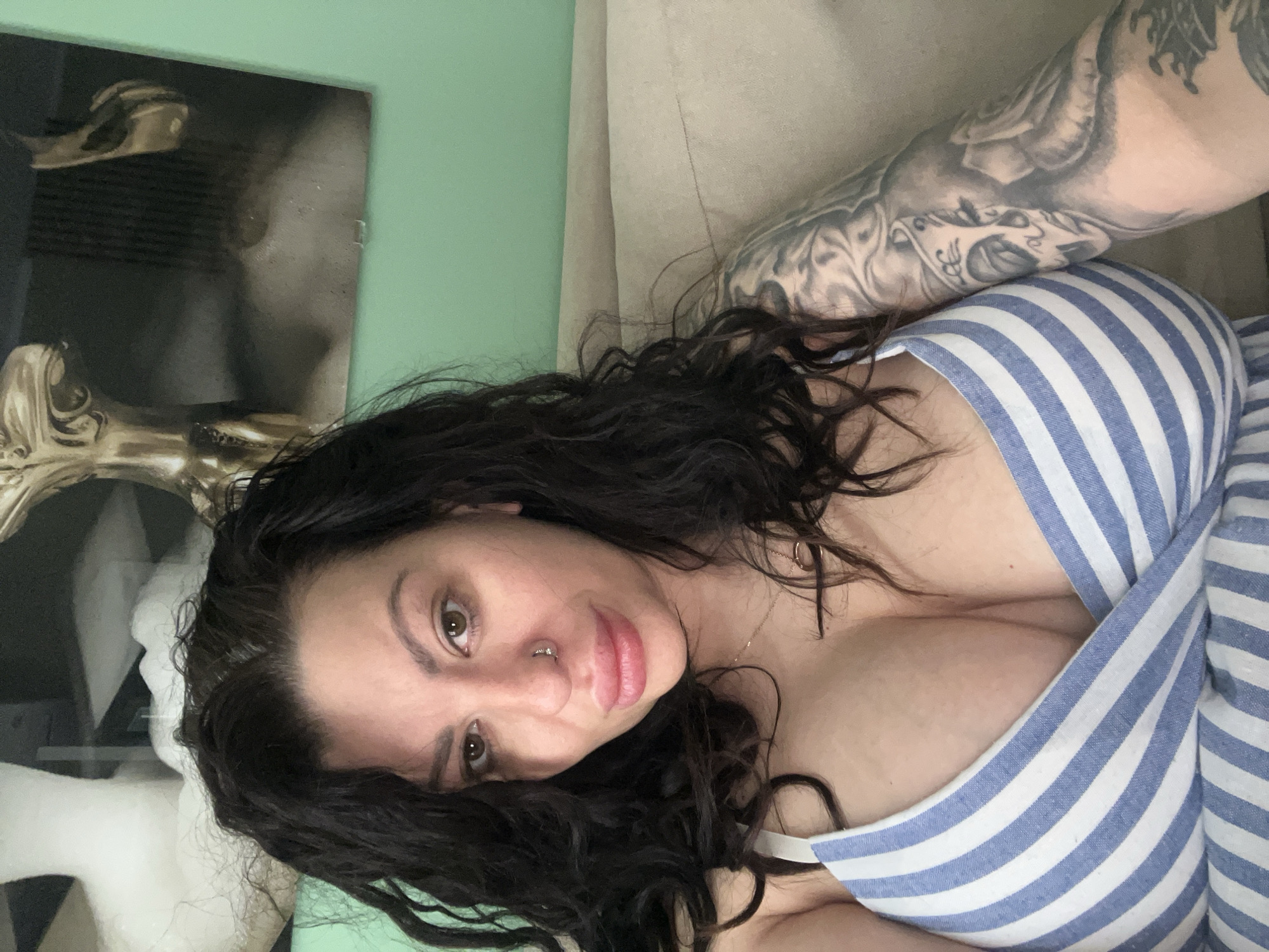 A woman with dark, wavy hair, fair skin, and a nose ring is lying down, partially visible in the image. She is wearing a blue and white striped garment. Her right arm is visible, featuring a large tattoo. The background includes a green wall, a beige sofa, and what appears to be a framed picture or artwork. The picture contains a figure and is placed against the green wall. The photo was not taken in Plovdiv, Bulgaria and there is no evidence to suggest it was.