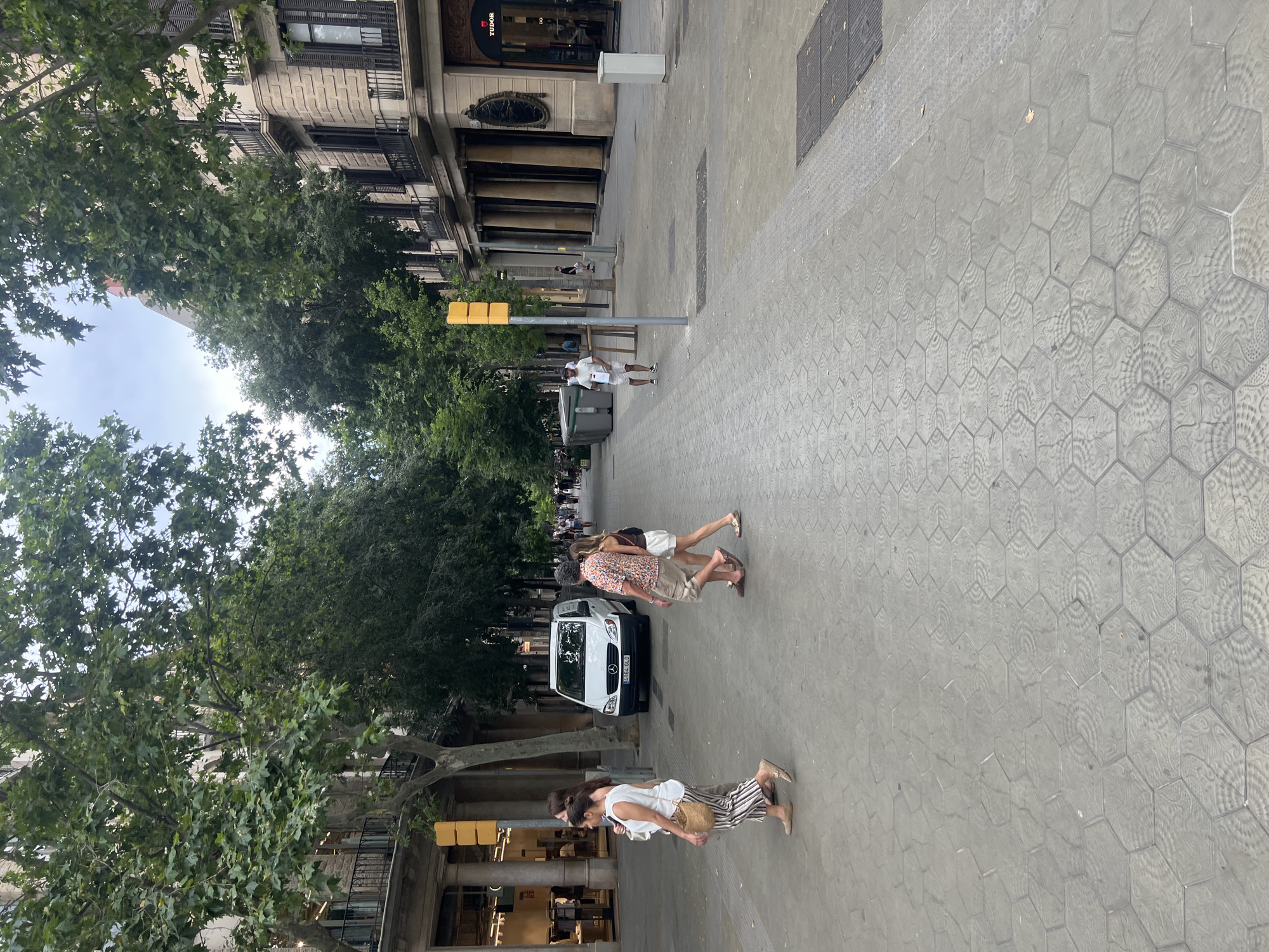 The image depicts a pedestrian walkway lined with trees and buildings in Barcelona, Spain. The walkway is paved with hexagonal tiles featuring an intricate pattern. Several people are walking on the sidewalk. In the foreground, a woman with striped pants and a white top walks from left to right, carrying a straw bag. Behind her, a couple walks in the same direction. Further down the street, a white van is parked. On the right side of the street, a building with columns and balconies is visible, housing shops including a Tudor watch store. The sky is overcast, and the overall atmosphere suggests a daytime scene.