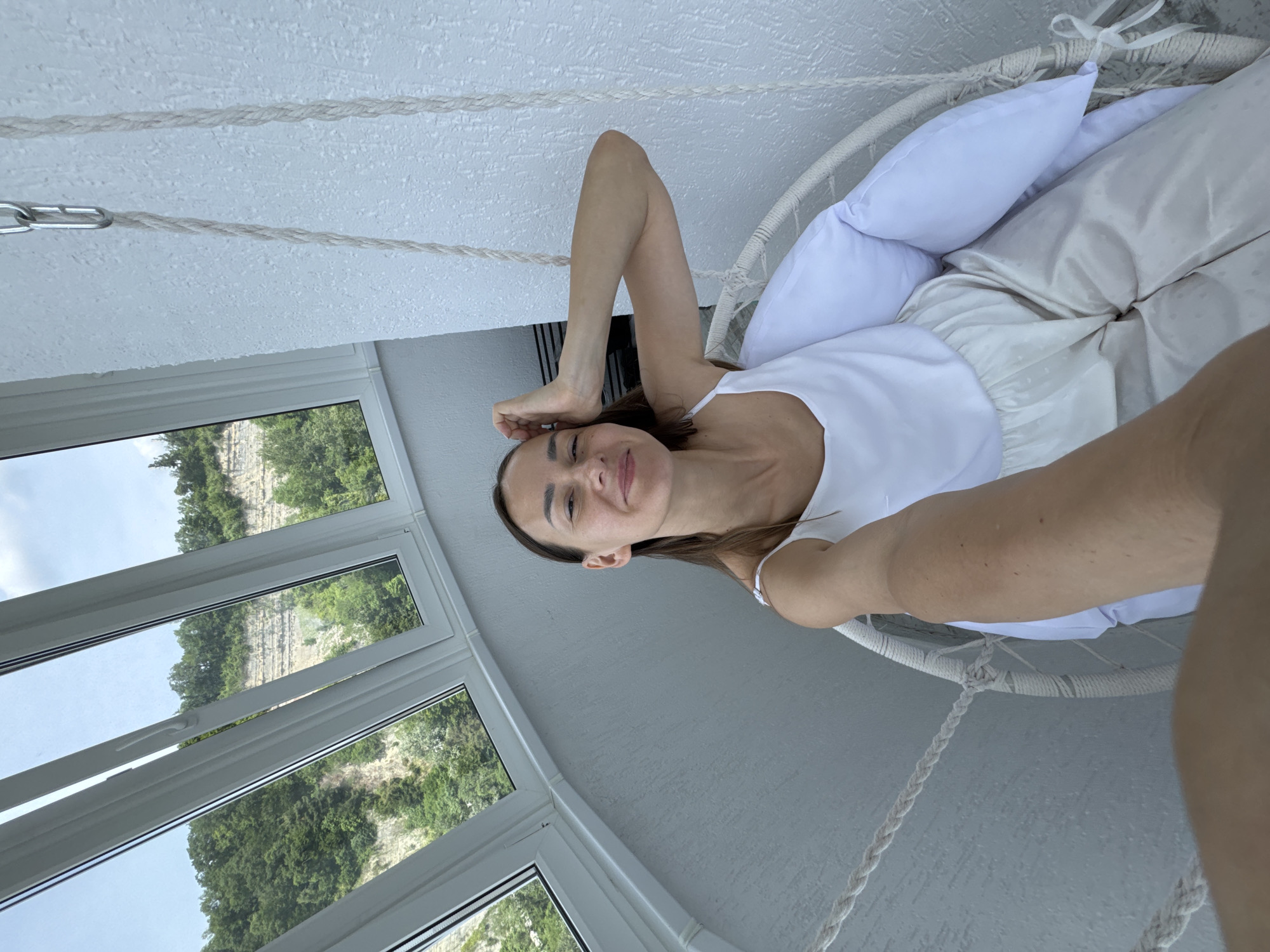 A woman is sitting in a white hanging chair on a balcony with a view of a green, rocky hillside and a cloudy sky through a large, angled window. The woman is wearing a white tank top and light-colored pants. She is smiling and has her hand on her head. The hanging chair is made of woven material and rope, with white cushions. The balcony wall is textured and white. The scene appears to be in a residential building in Sochi, Russia, given the lush vegetation and rocky cliffs visible outside.